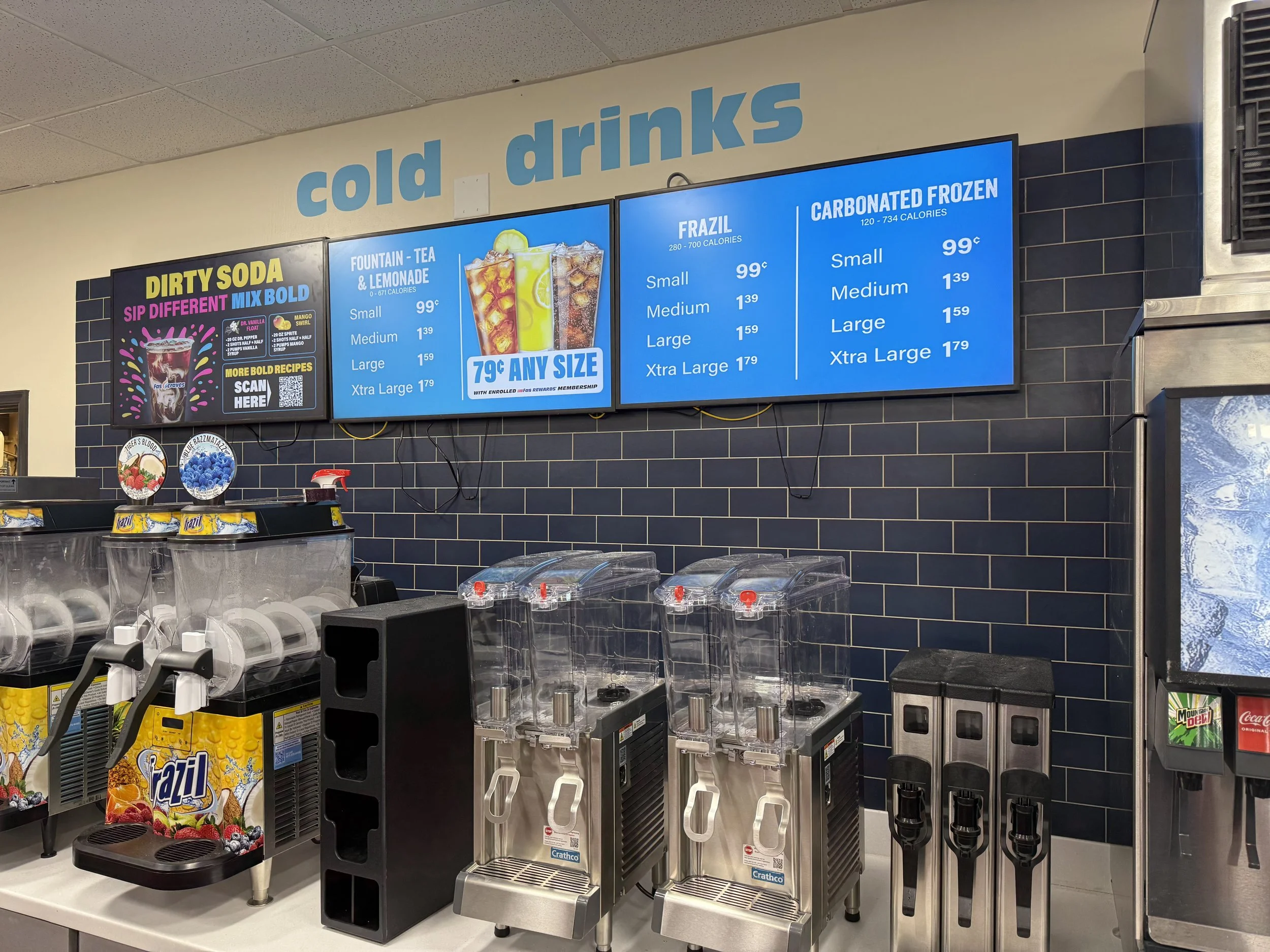 Digital menu display showing prices for frozen drinks like Frazil and Carbonated Frozen in small, medium, large, and extra large sizes with colorful signage and beverage dispensers below.