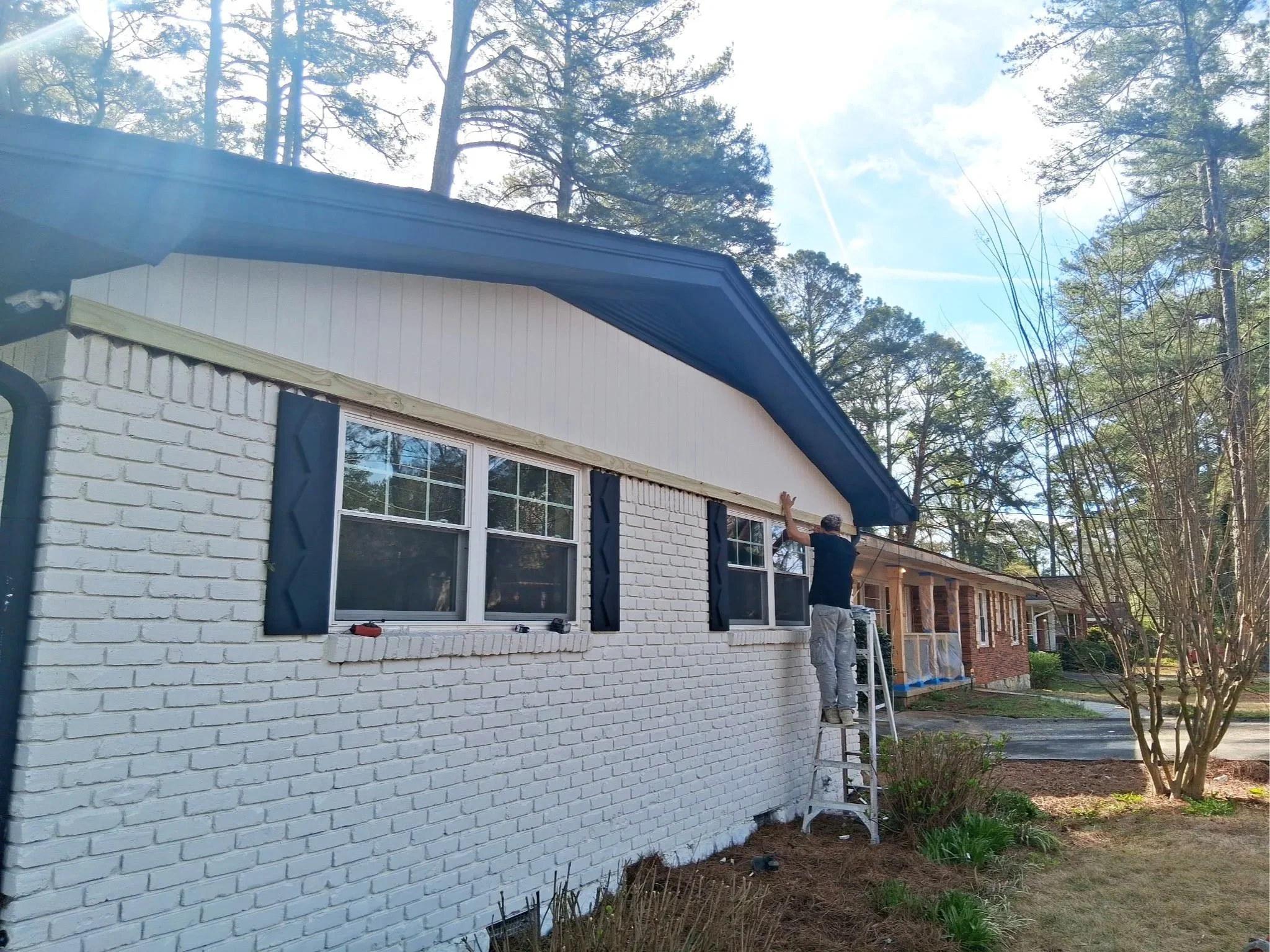 A person on a ladder painting the exterior of a residential house's trim white, with the house having white brick walls, black shutters, and large windows, surrounded by trees and a garden.