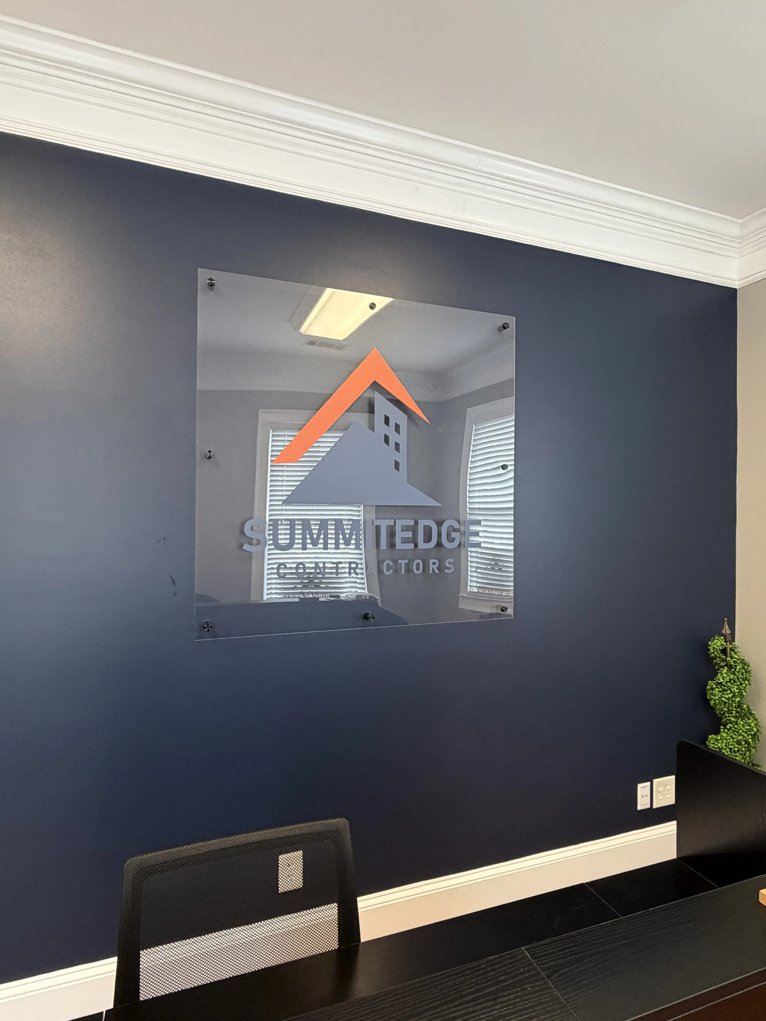 Office conference room with a navy blue accent wall, a glass wall sign for Summit Edge Contractors, and a black conference table with a mesh chair. There is artwork of a green twisted topiary on a stand in the corner and window blinds reflected in the glass sign.