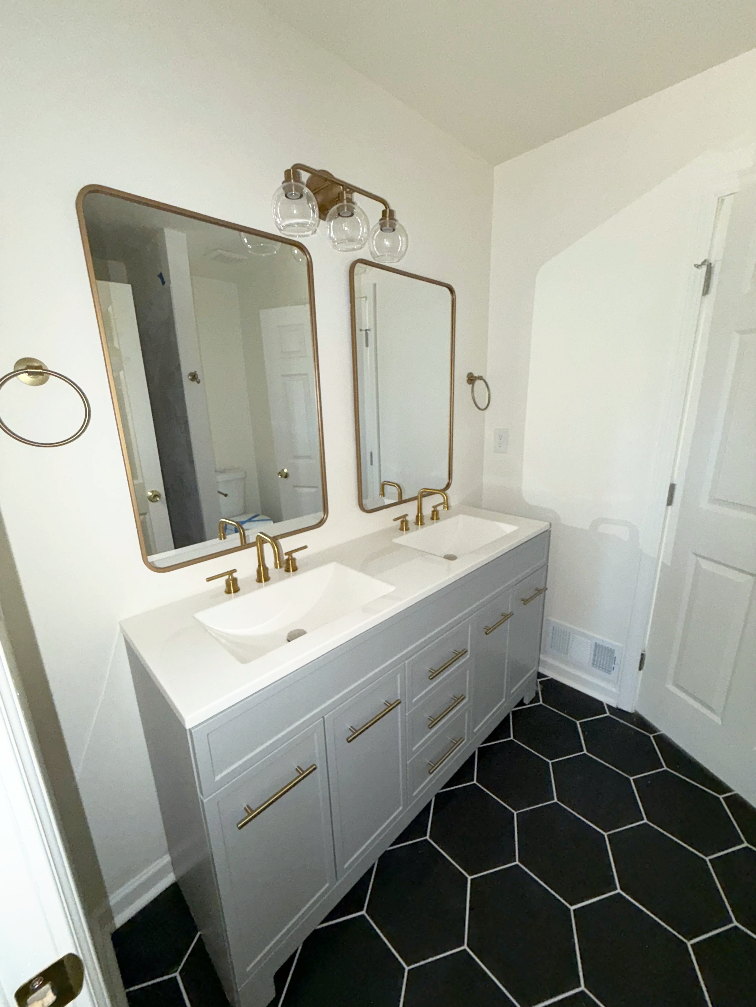 Double vanity bathroom with two white vessel sinks, gold faucets, large rectangular mirrors with gold trim, black hexagonal floor tiles, white walls, and two gold towel rings.