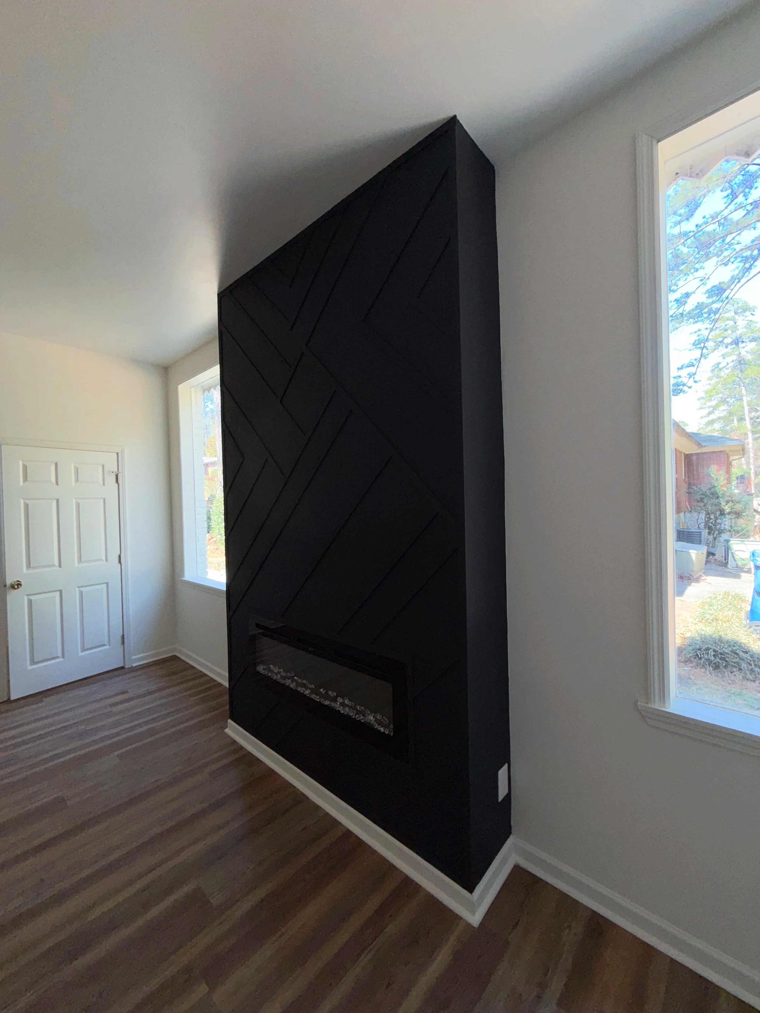 Modern black fireplace with geometric wood panel design in a bright living room with white walls, hardwood floors, and large windows.