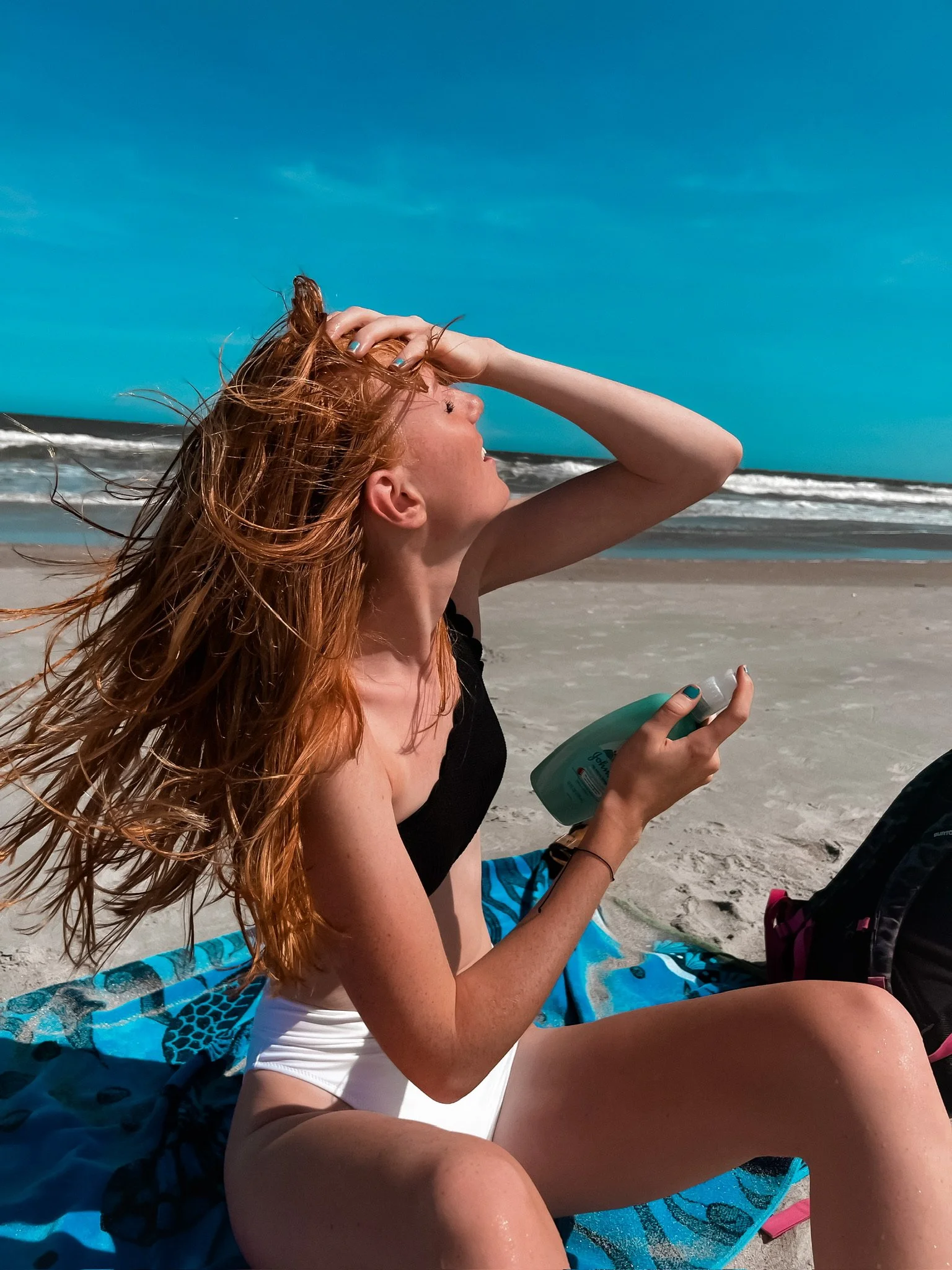 Young woman sitting on a beach towel applying sunscreen, with her hair blowing in the wind, on a sunny beach with the ocean waves in the background.