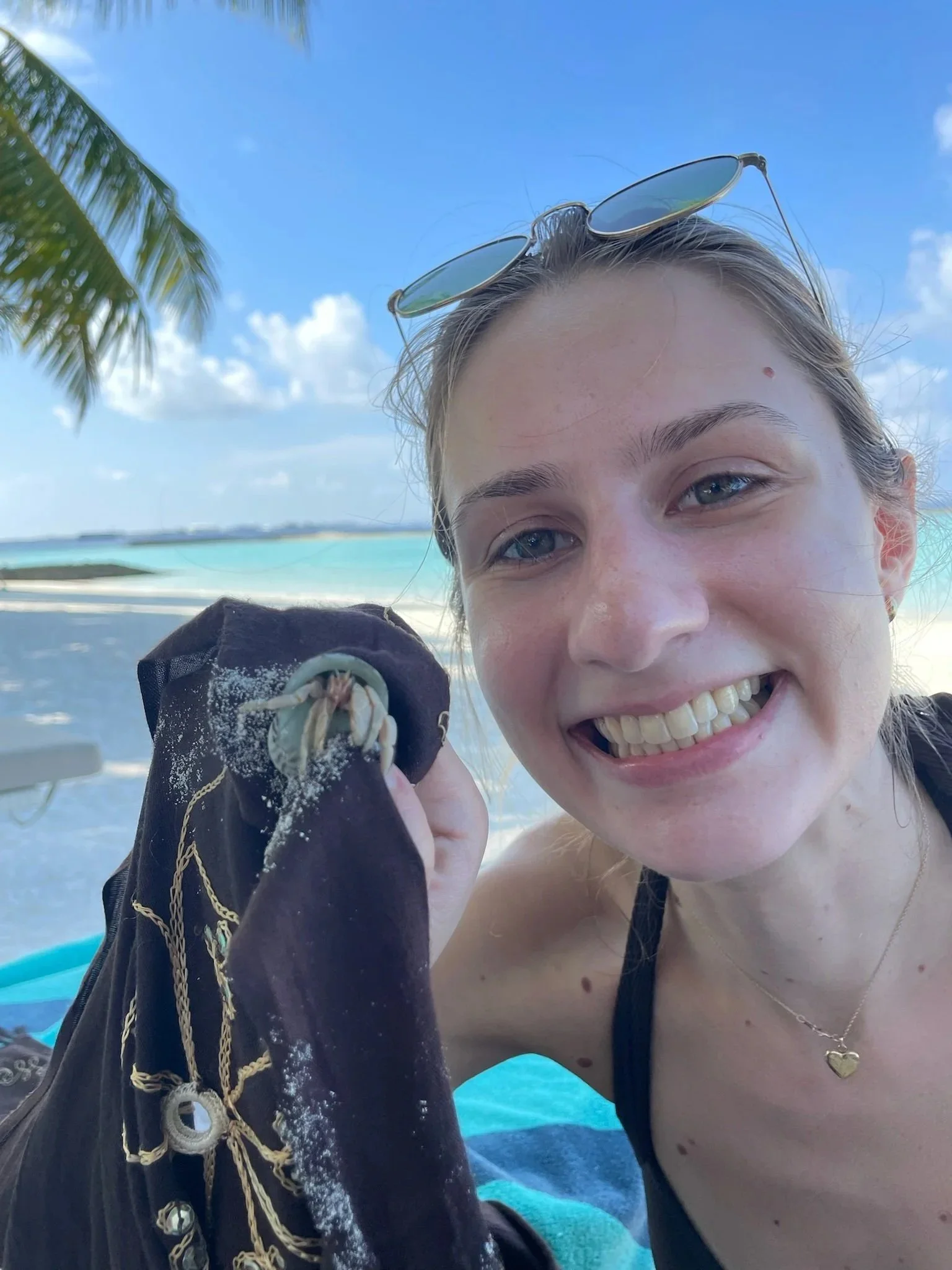 A woman smiling at the camera, holding a seashell by the beach with clear blue ocean and sky in the background.