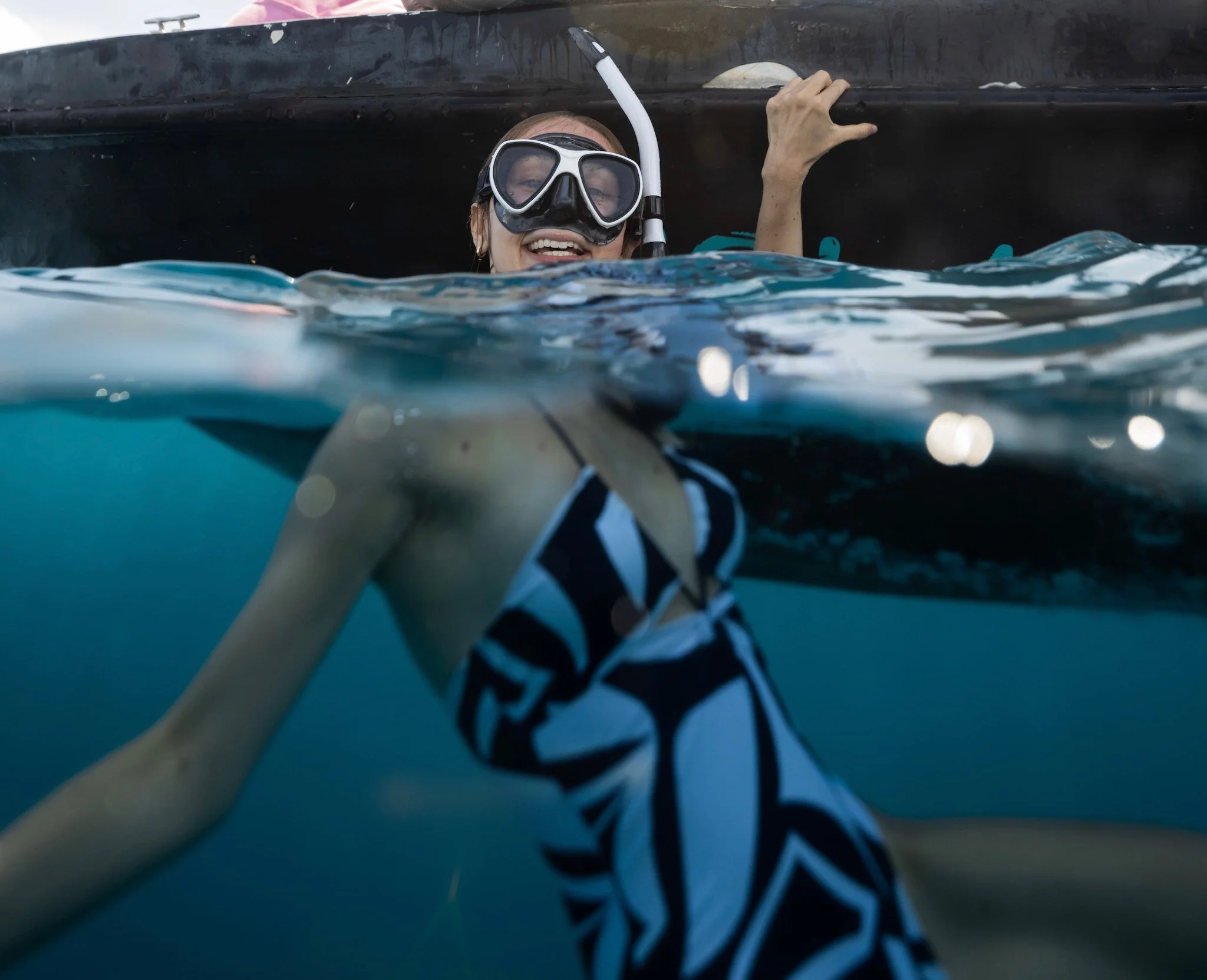 Woman wearing snorkeling gear, smiling, half above water and half submerged, in an outdoor water setting.
