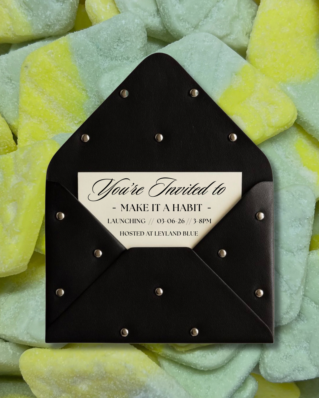 Invitation card inside a black envelope with silver studs, placed on a pile of pastel green and yellow candies.