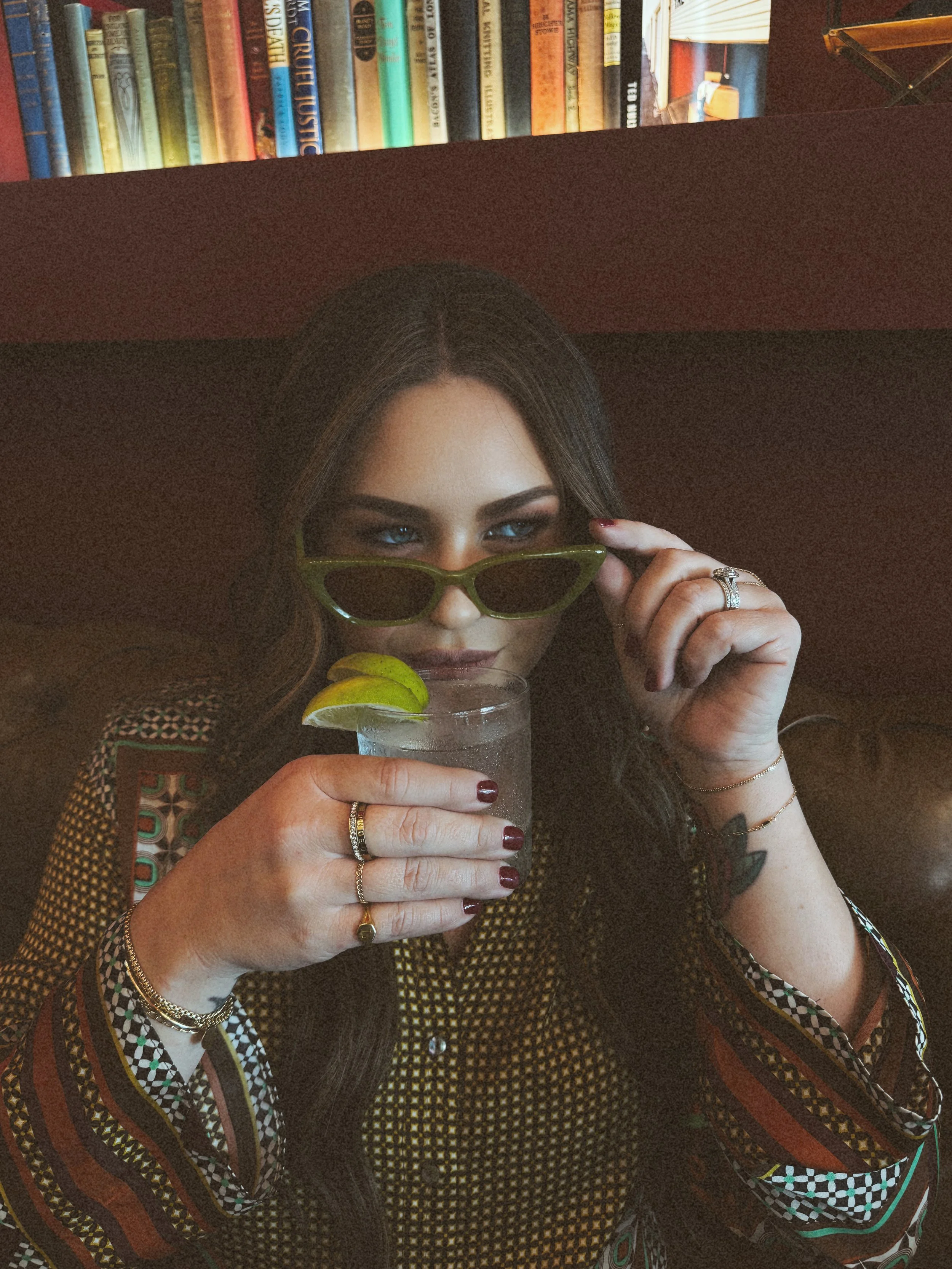 A woman holding a glass of drink with lime wedge, adjusting green sunglasses, sitting in a cozy environment with bookshelf in background.