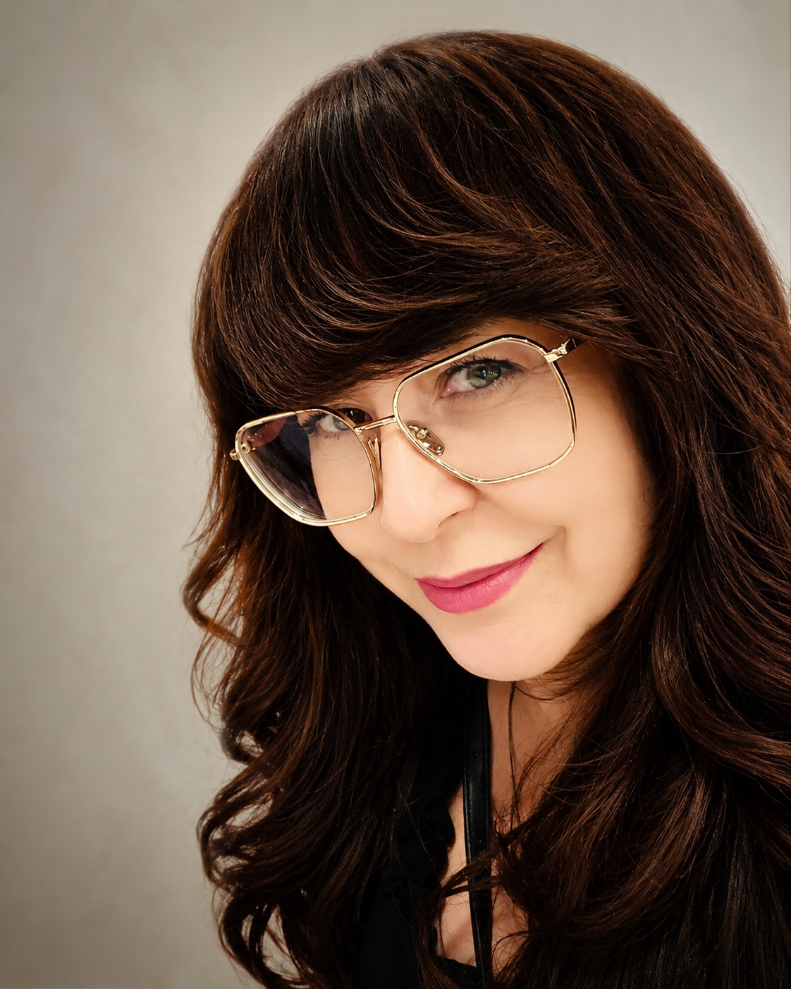 Close-up of a woman with wavy brown hair, wearing large gold-rimmed glasses, smiling at the camera, against a light gray background.