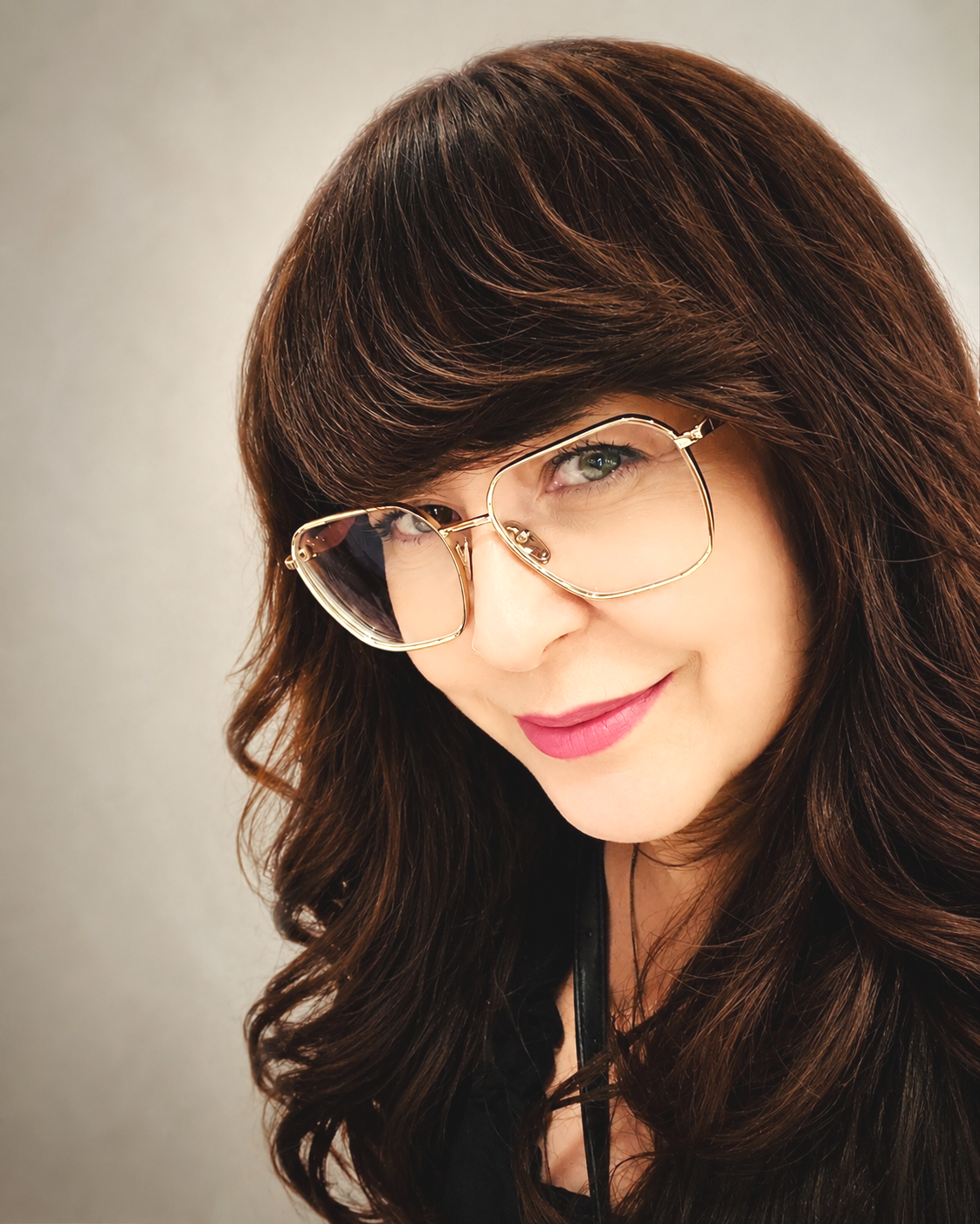 A woman with long brown hair and light skin wearing large gold-rimmed glasses, smiling slightly, against a plain light background.