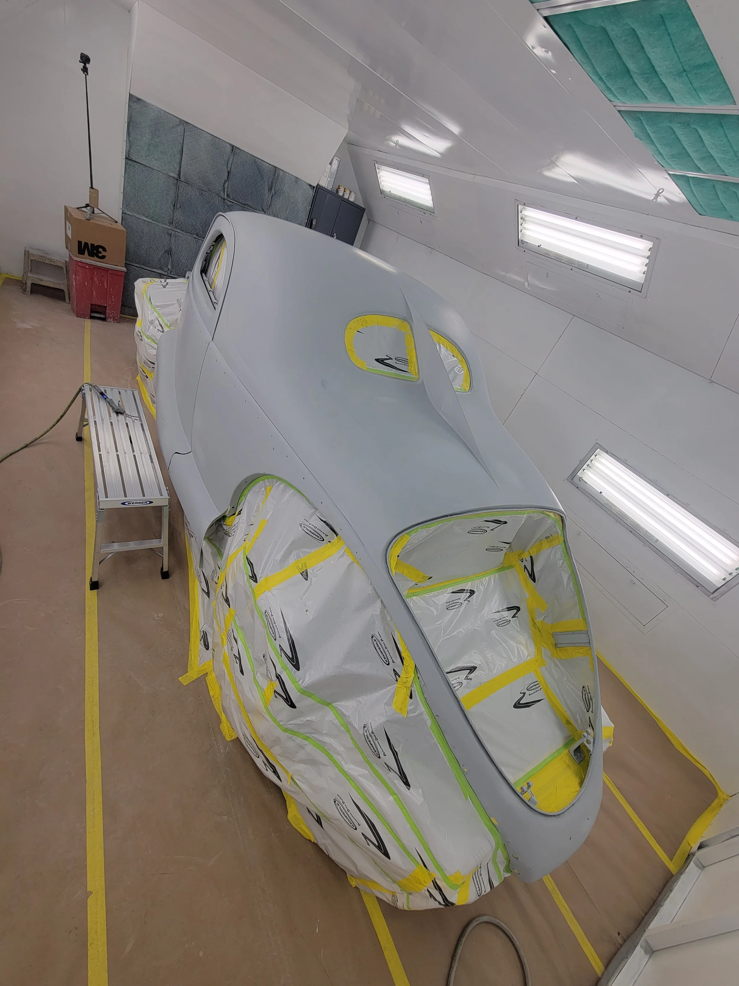 A classic car being painted in a spray booth. The car's body is primed and masked, with protective plastic and yellow masking tape covering certain areas.
