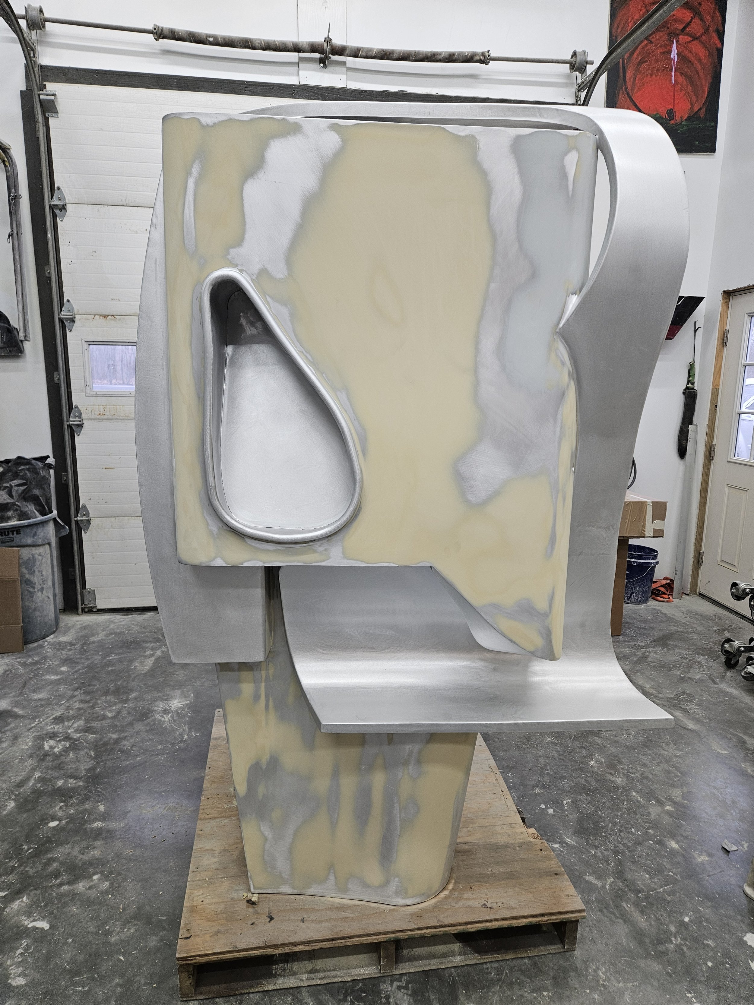 Unfinished metallic sculpture in a workshop, resting on a wooden pallet, with various tools and a painting on the wall in the background.