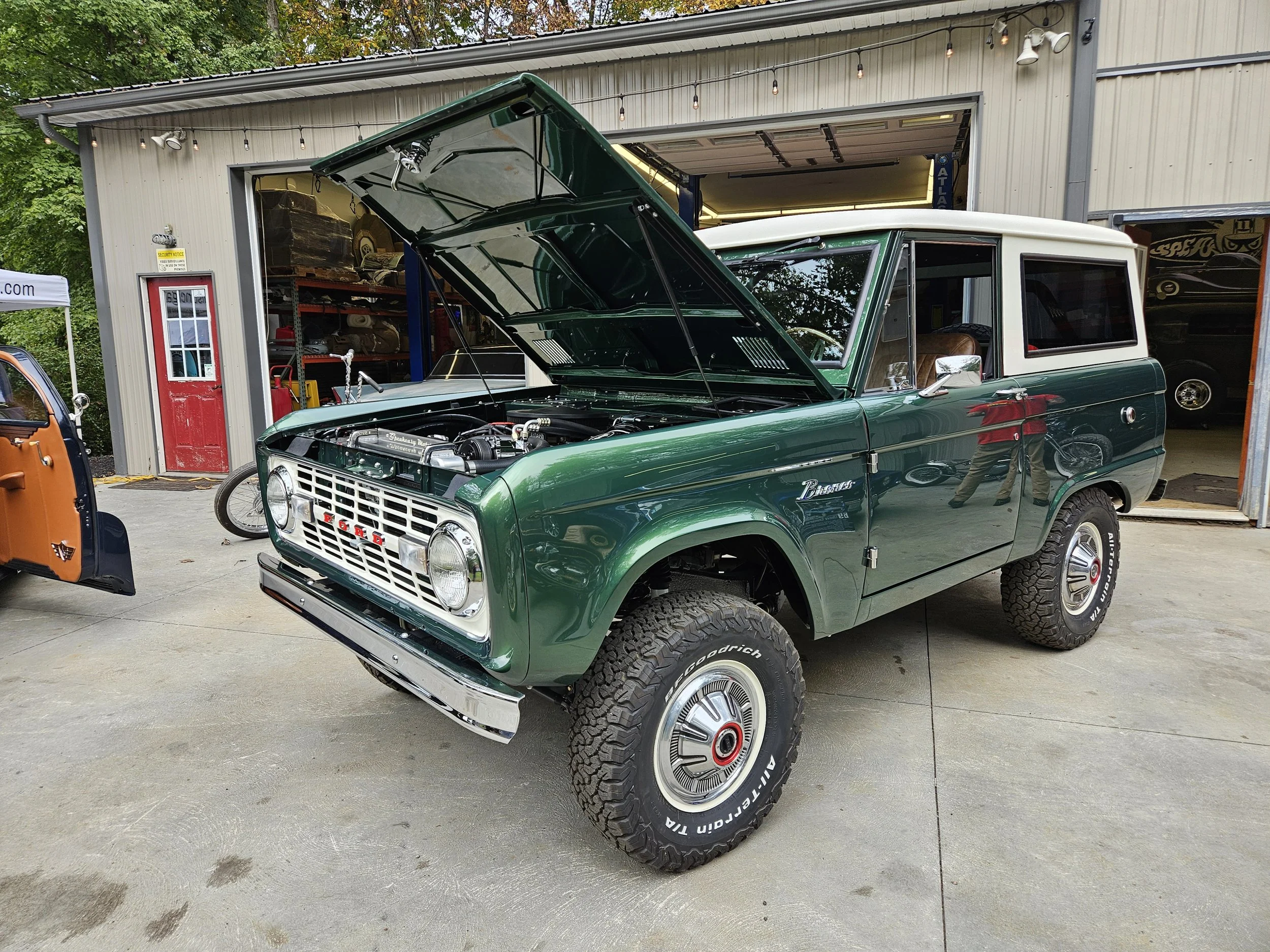 A vintage green and white SUV with its hood open, parked outside a garage. The vehicle has large off-road tires and a classic design.