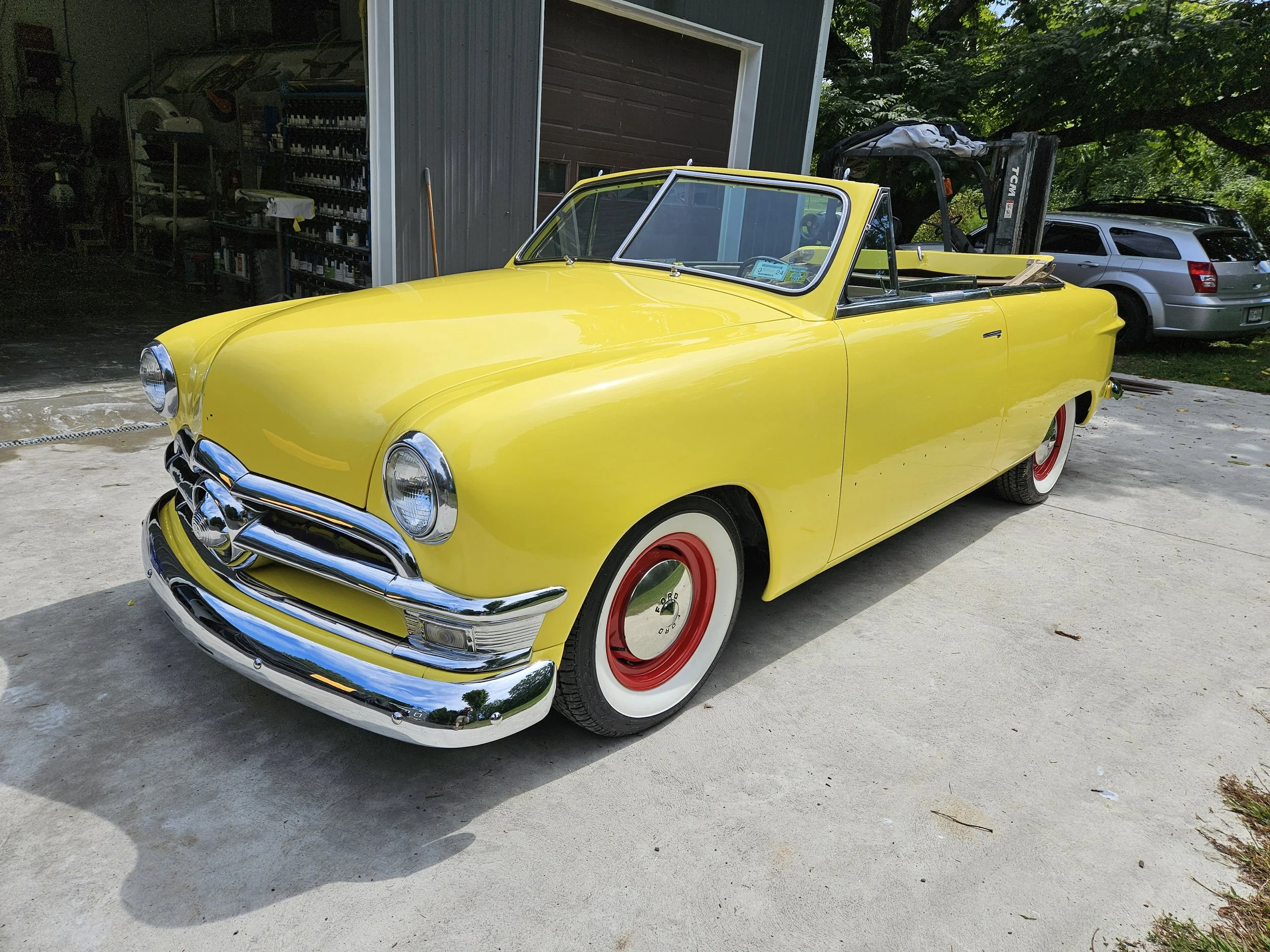 A bright yellow vintage convertible car with whitewall tires, chrome bumpers, and red hub caps, parked on a concrete driveway near a garage.