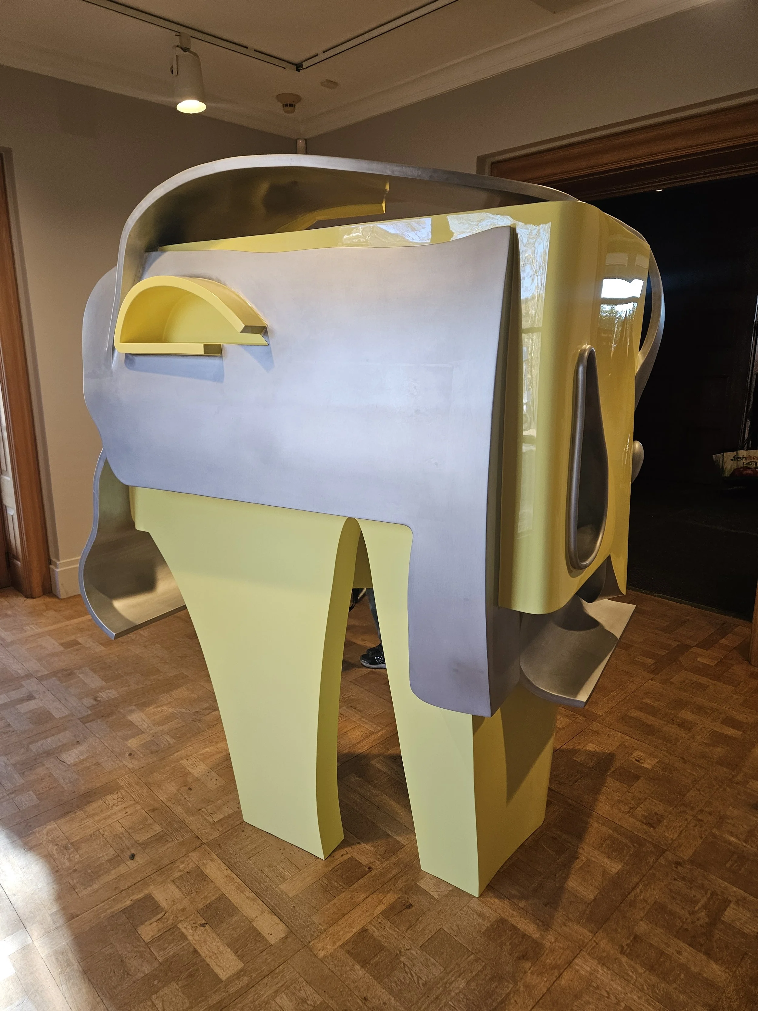 A modern, abstract art sculpture resembling a stylized robot or animal, with yellow and metallic silver components, placed on a wooden floor in an indoor setting.