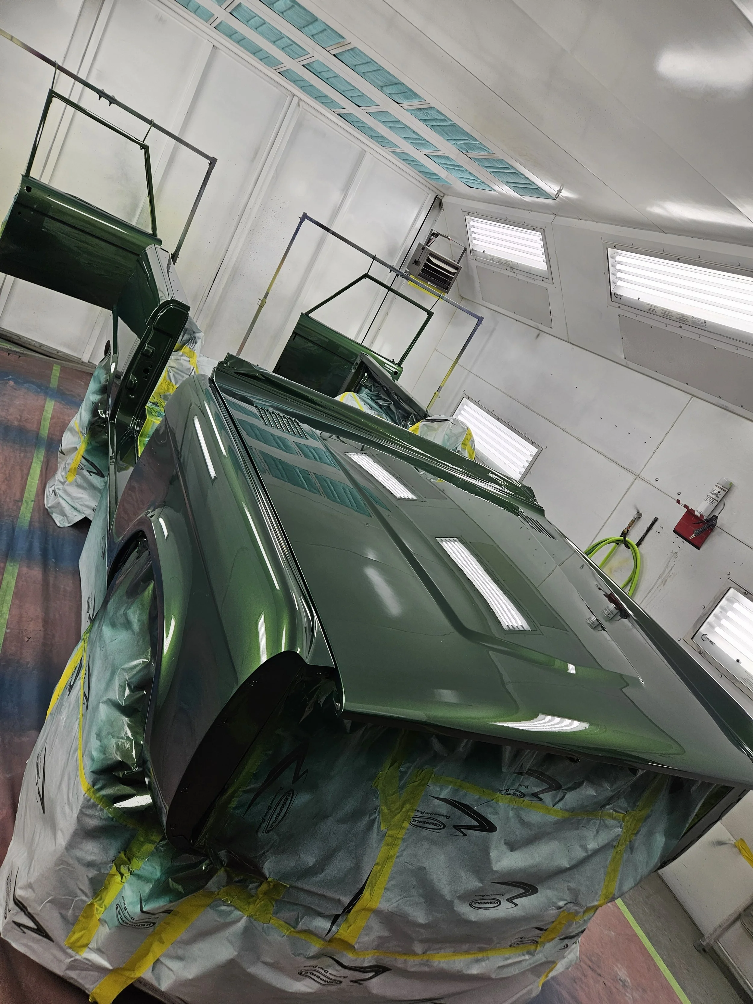 A green car in a spray booth being painted, covered with masking paper around the wheel wells and edges for protection during painting.