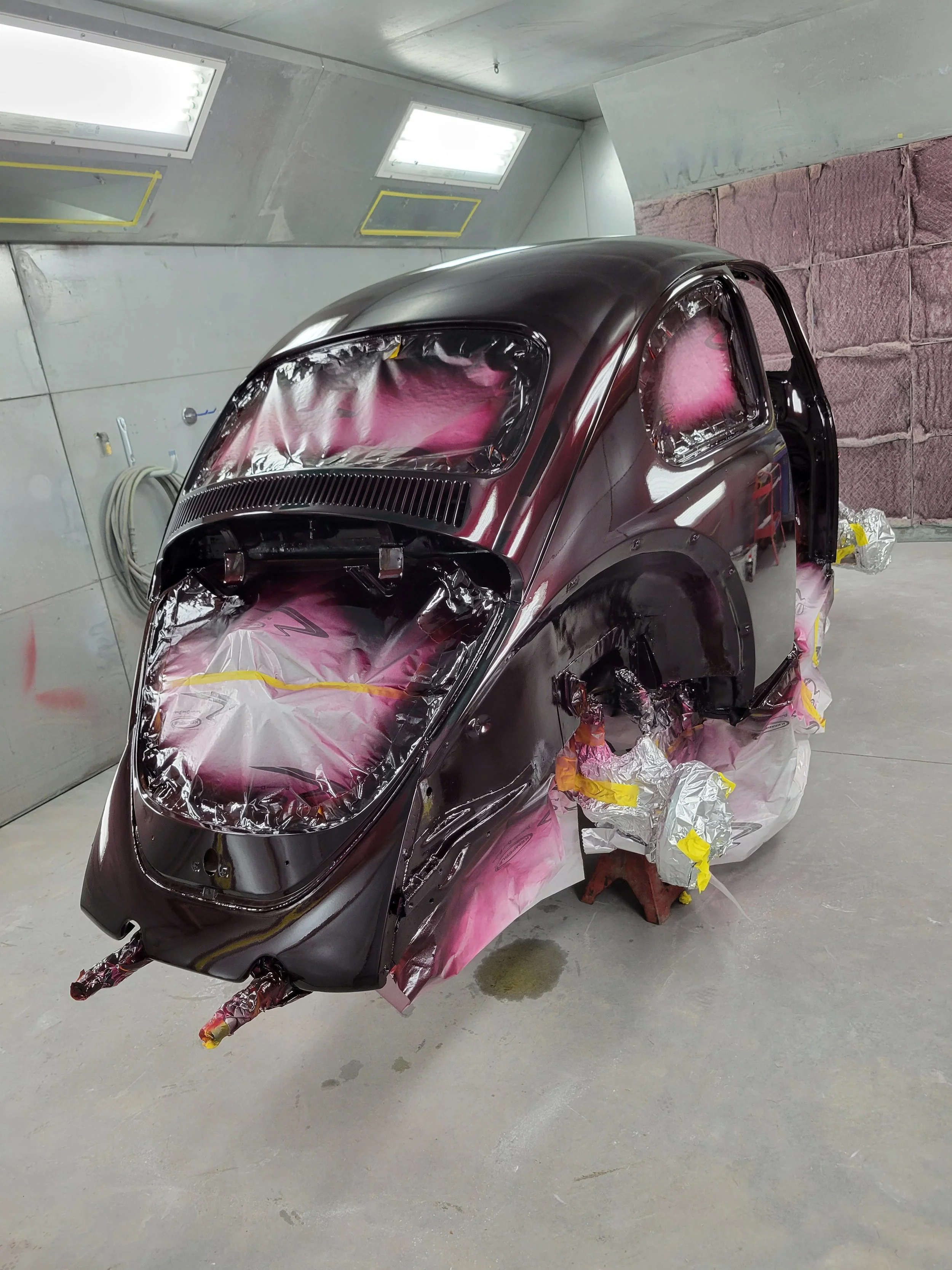 Black vintage car body in a paint booth, masked with protective tape and plastic for painting, elevated on supports, with plastic sheeting and paper covering parts of the car.
