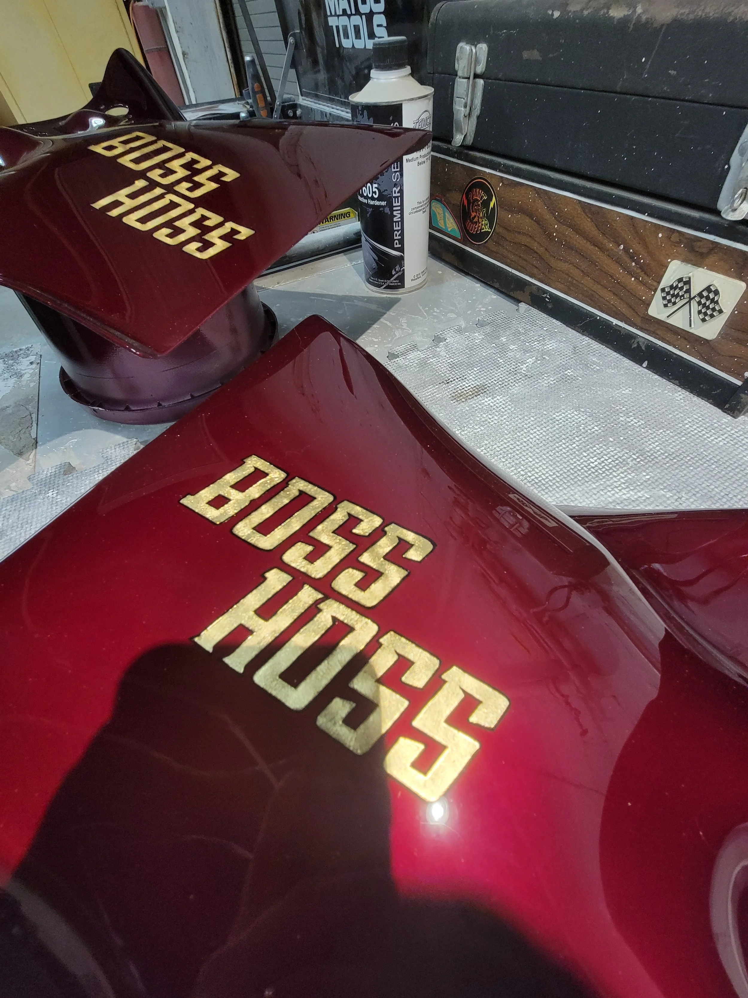 Red motorcycle with gold lettering that says 'BOSS HOSS' on the fairing, inside a workshop with various tools and supplies, including a can of chain lube and stickers on the wall.