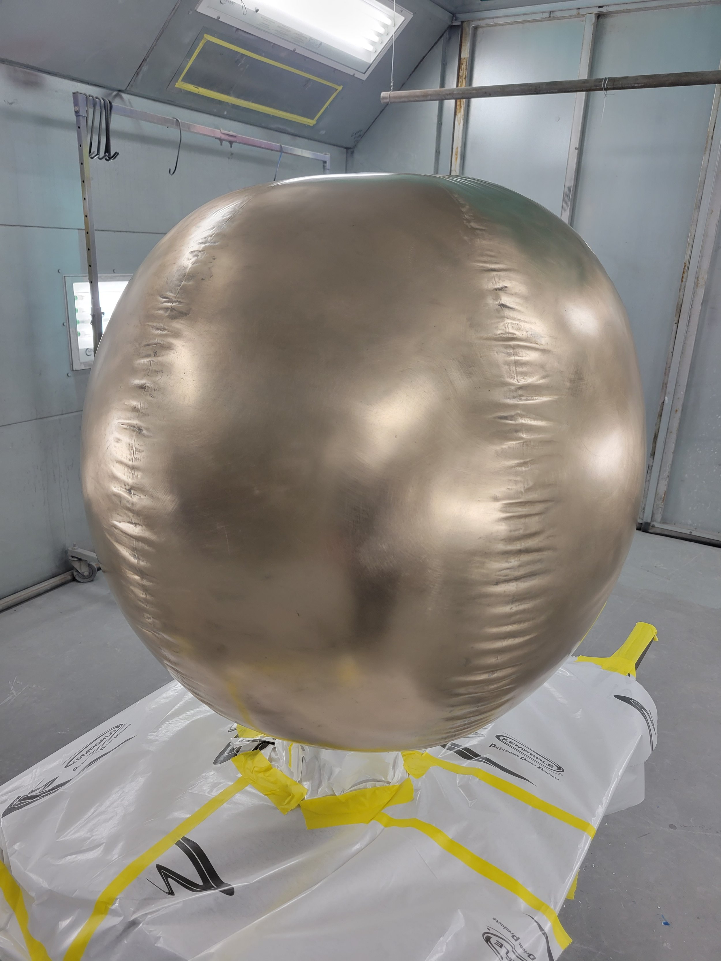 Large metallic sphere in a manufacturing or industrial setting, positioned on a wrapped pallet in a painted metal room.