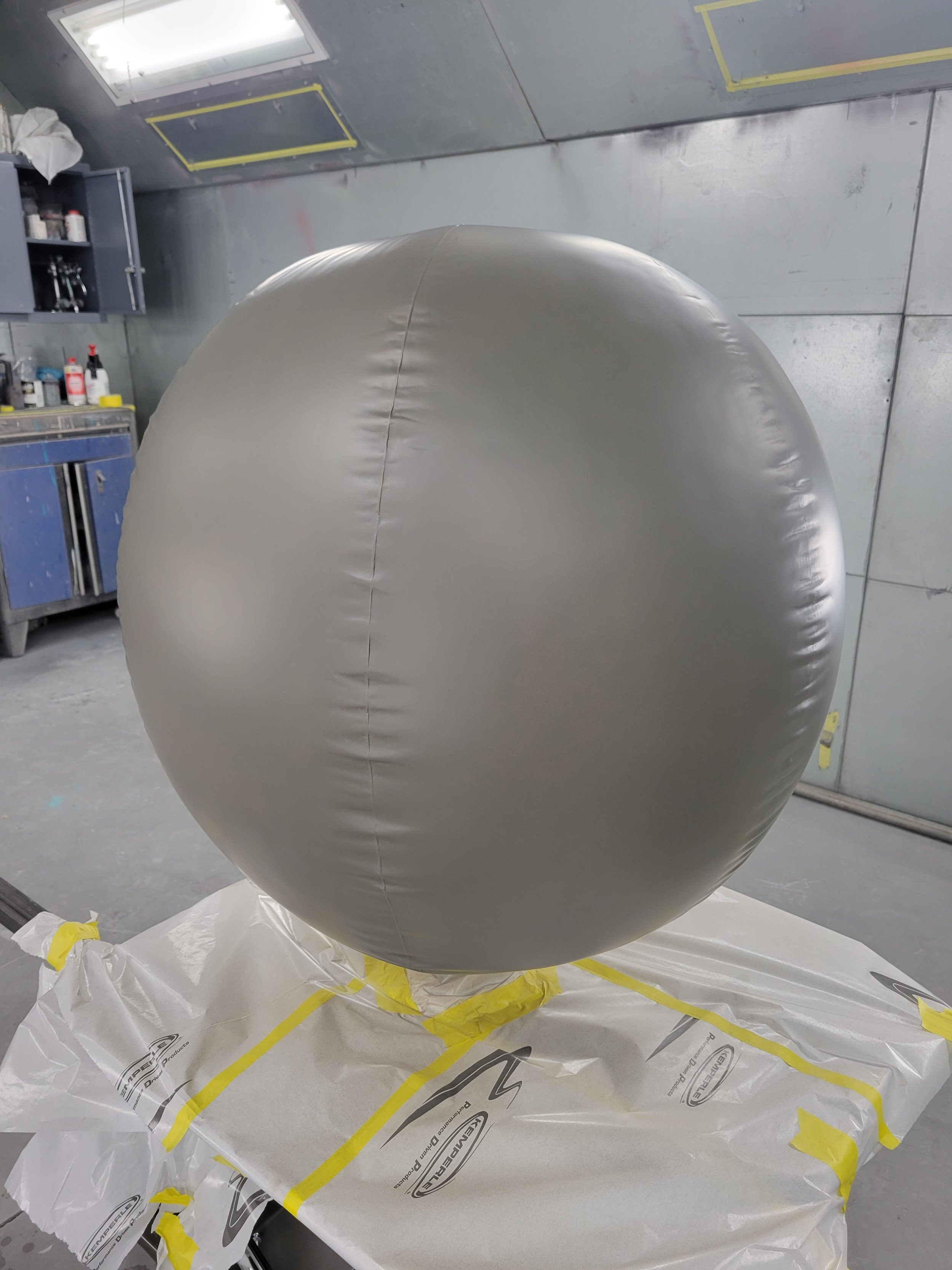 Large round object with a shiny, smooth surface, placed on a table covered with protective paper, in an industrial workspace with storage cabinets and tools in the background.