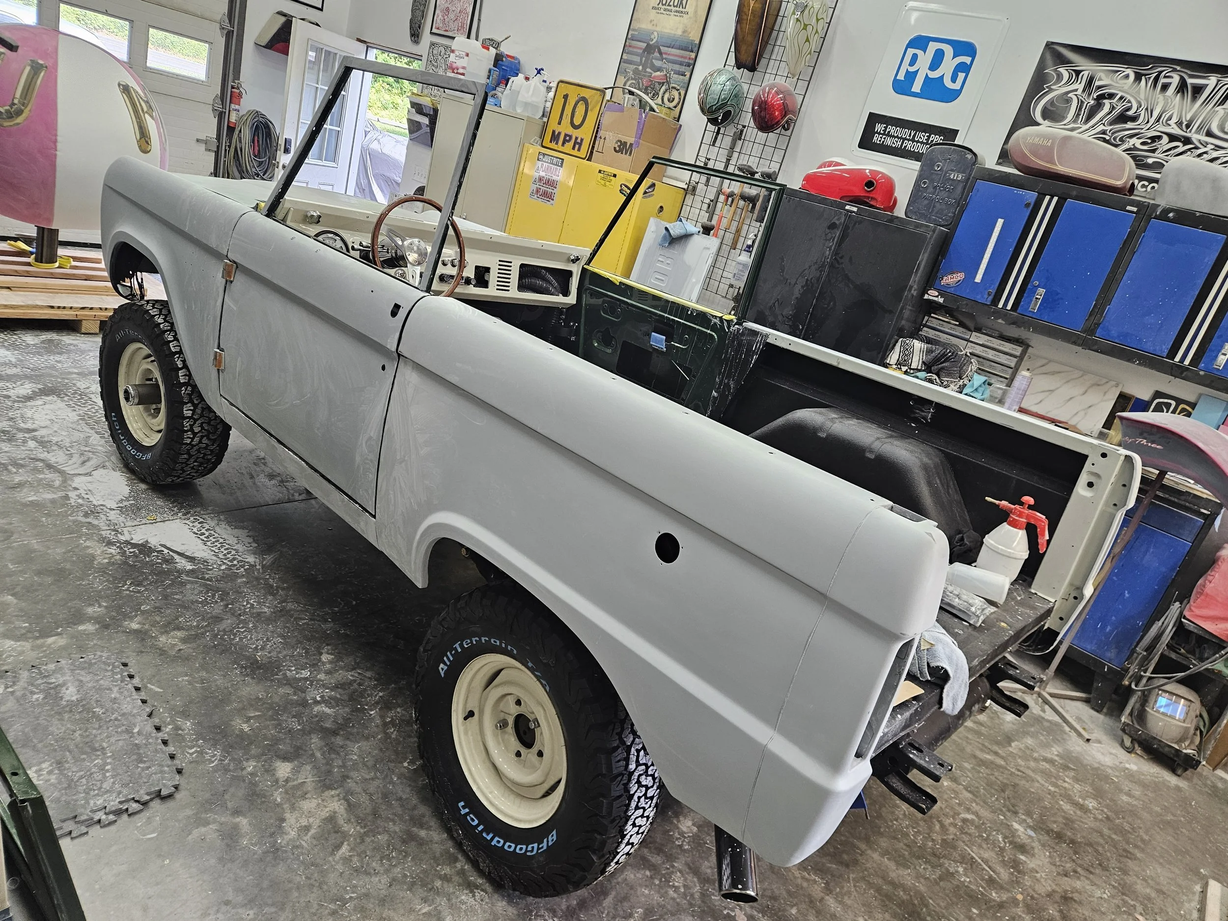 A partially assembled vintage pickup truck in a garage, with the bed and rear panels cropped out. The vehicle has black tires, a gray body, and a beige steel wheel. The interior shows a brown steering wheel and minimal dashboard. Garage shelves in th