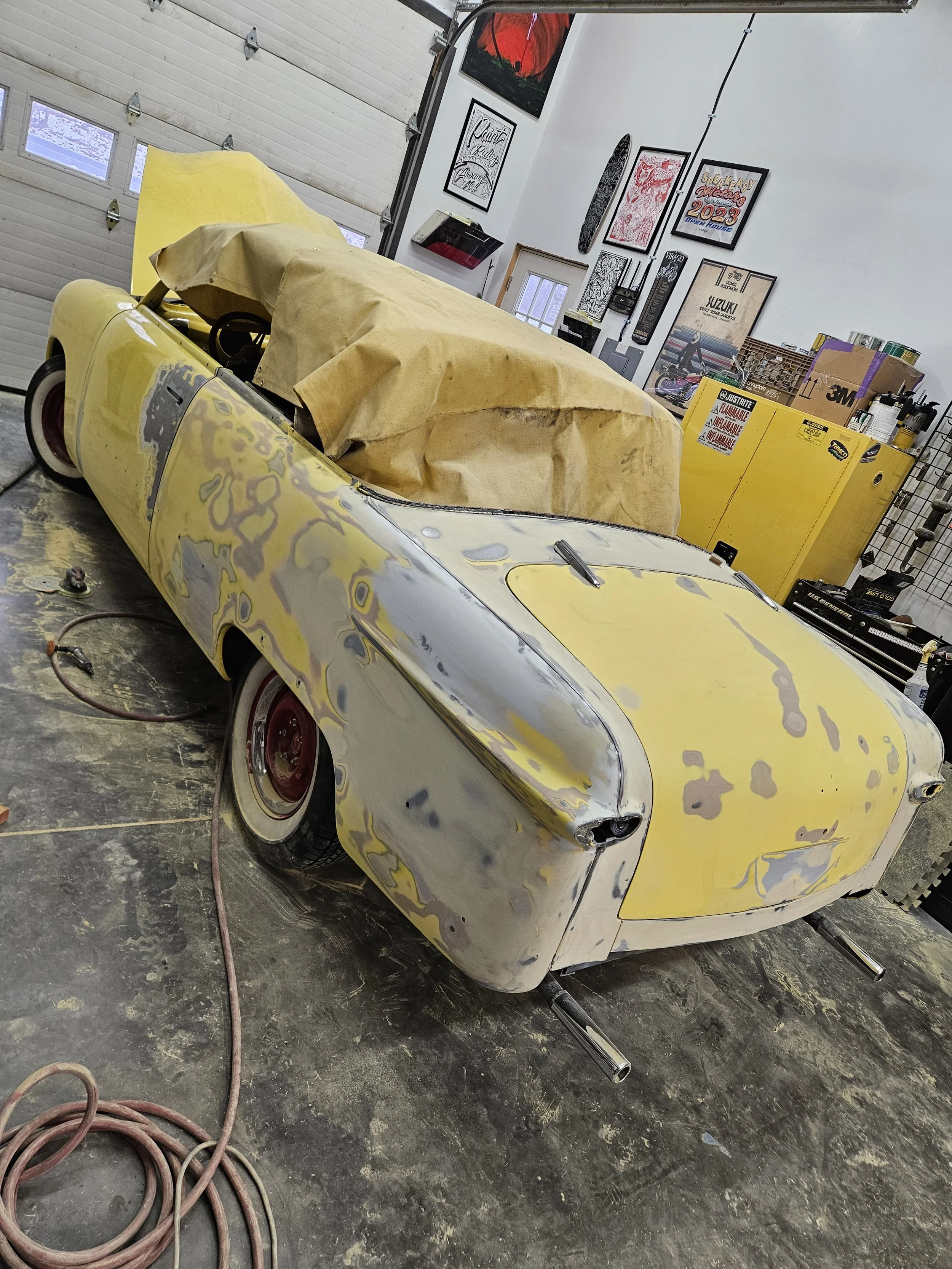 A vintage yellow and gray convertible car undergoing restoration in a garage, with parts masked off and a paper covering the interior, surrounded by tools and car supplies.