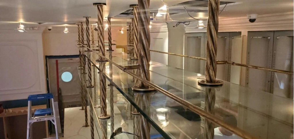 Interior of a building under construction with twisted metal posts, glass railing, ladder, and construction materials visible.