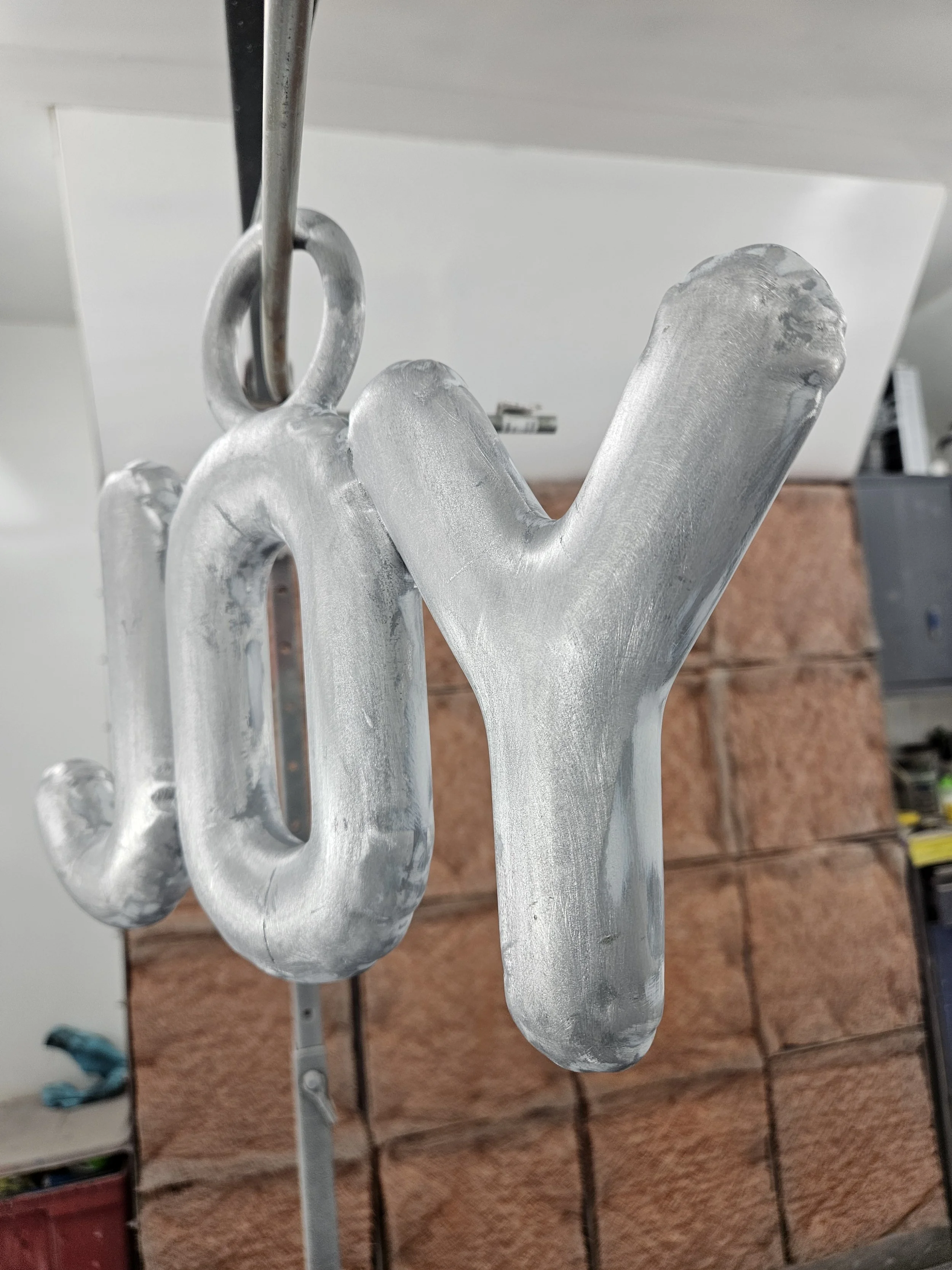 Close-up of a metallic, three-dimensional sculpture of the word 'joy' hanging from a hook.
