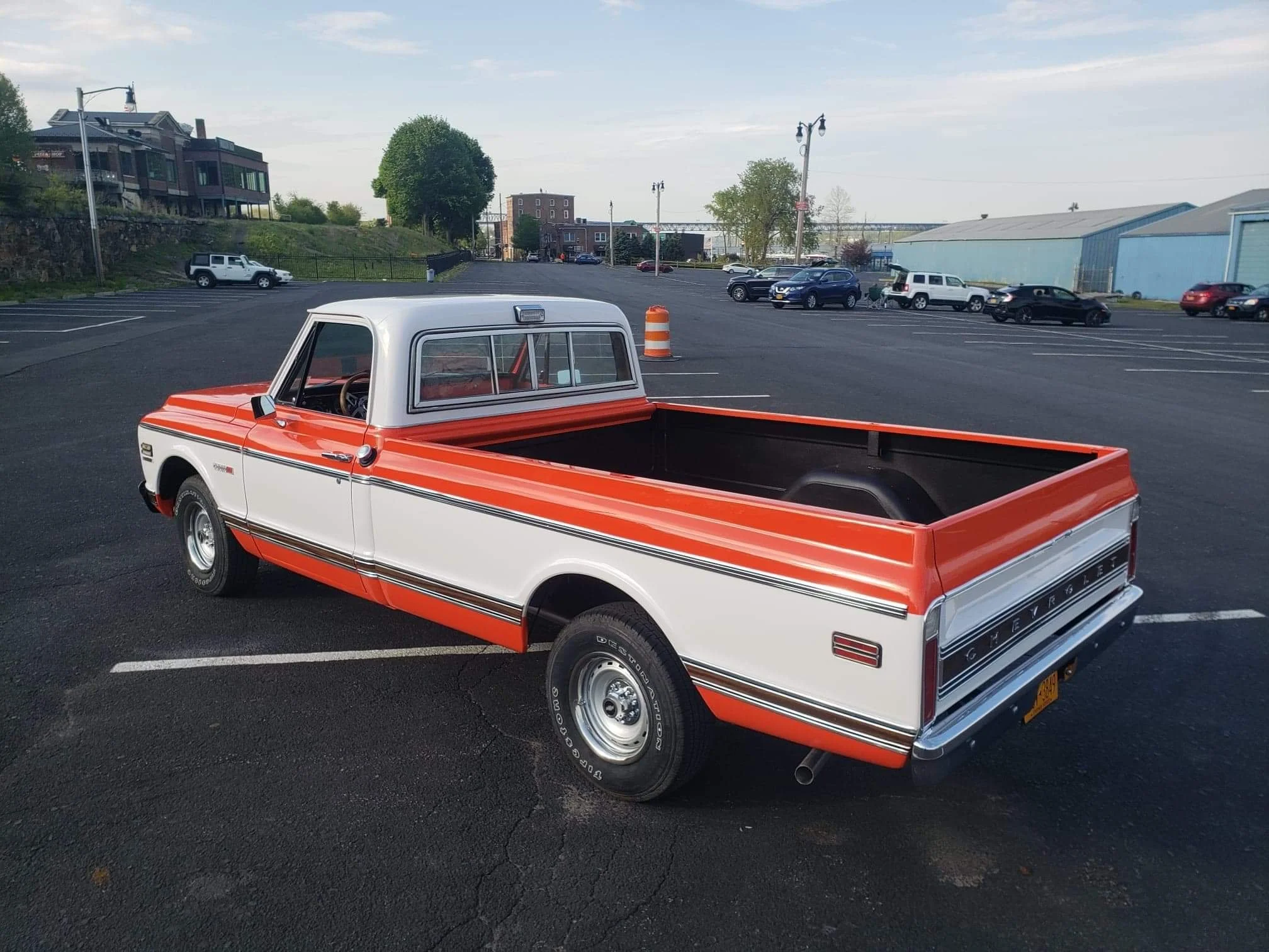 A vintage Chevrolet pickup truck with a red and white color scheme is parked in an open parking lot. The truck has a classic design with an empty bed, and other cars and buildings are visible in the background under a partly cloudy sky.