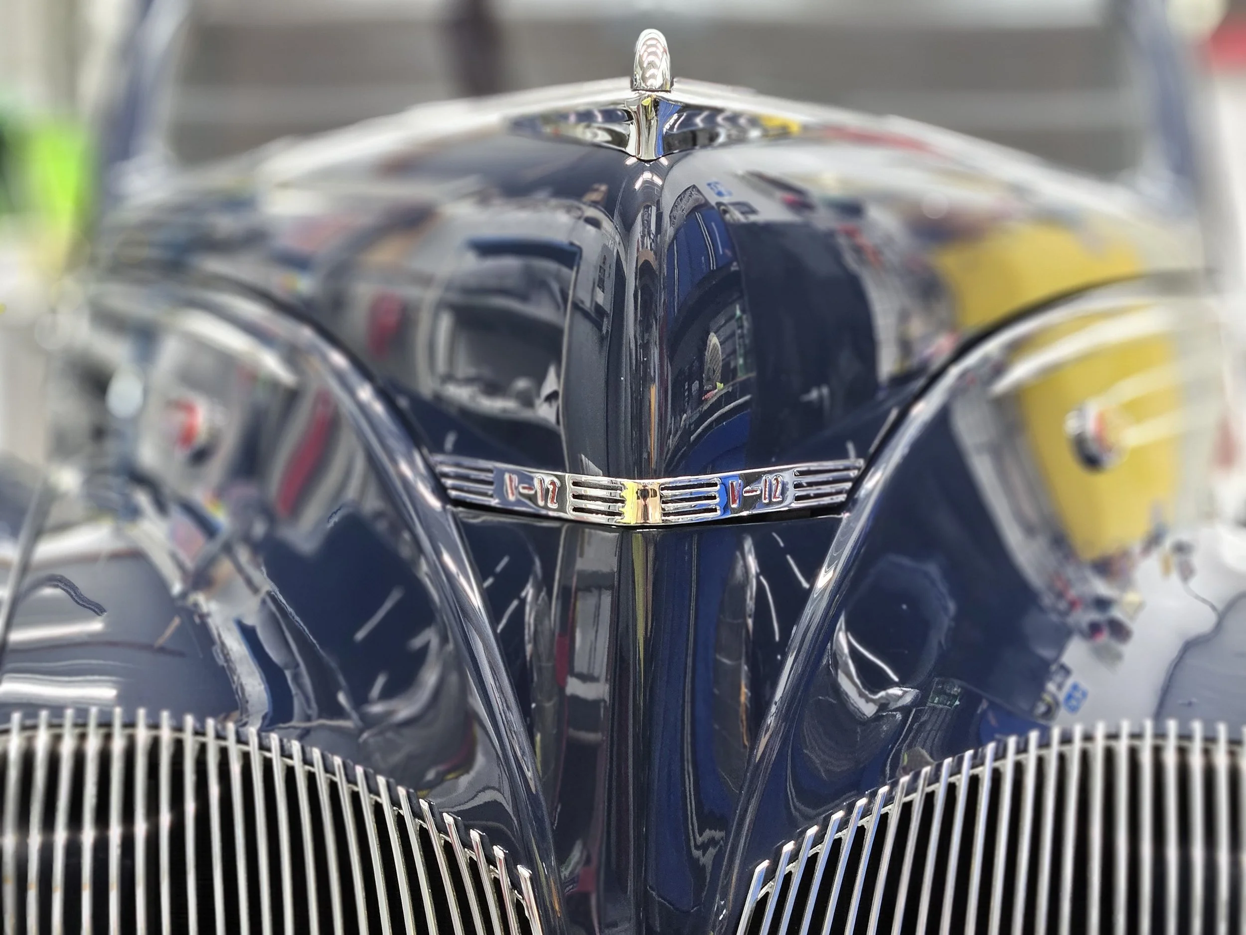 Close-up of a vintage blue car's front grille and hood ornament, with reflections visible on the shiny metal surface.