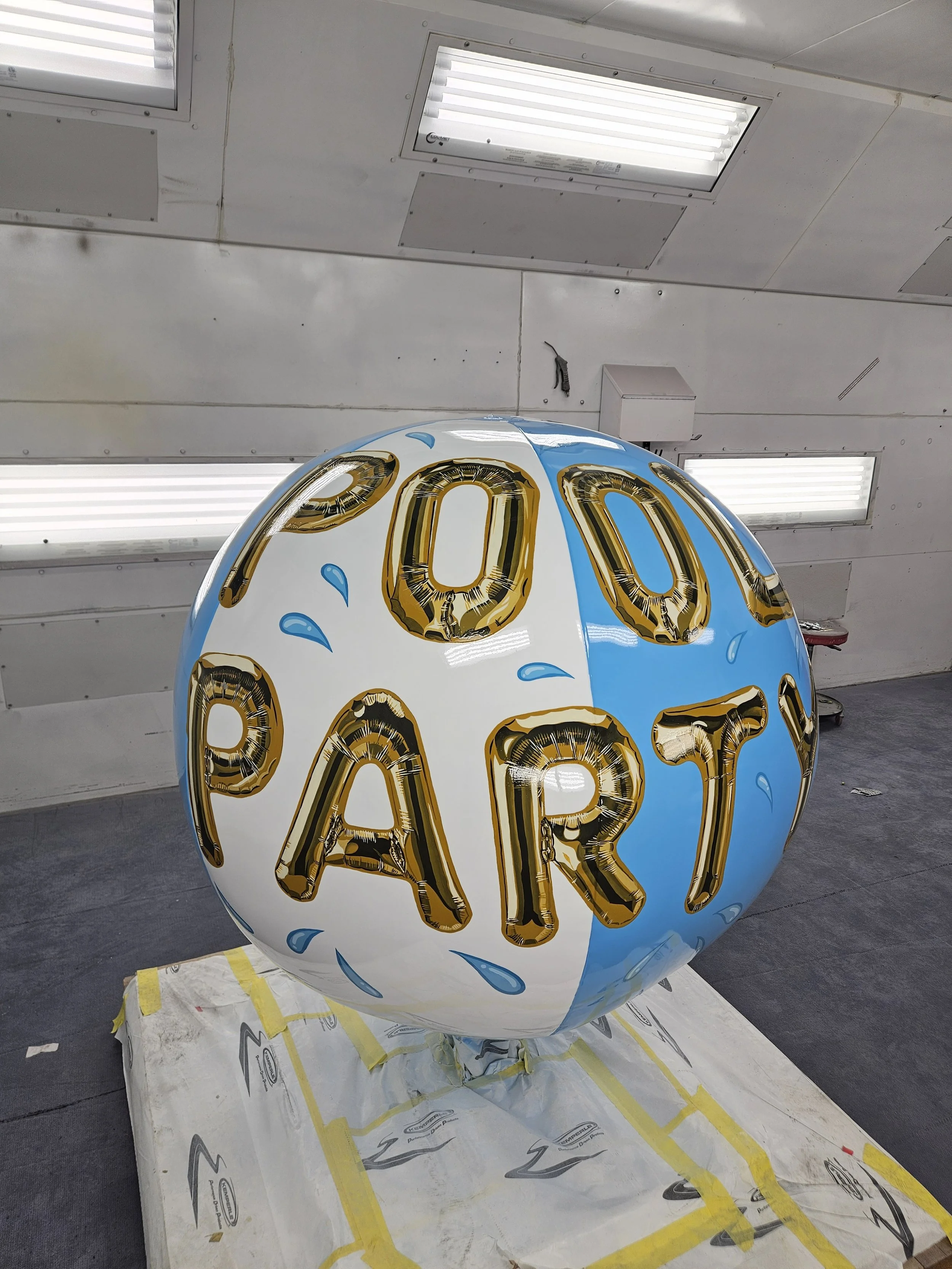 A large spherical balloon with the words 'POOL PARTY' in gold foil letters, partly blue and white, placed on a white tarp in an indoor space with fluorescent lights.