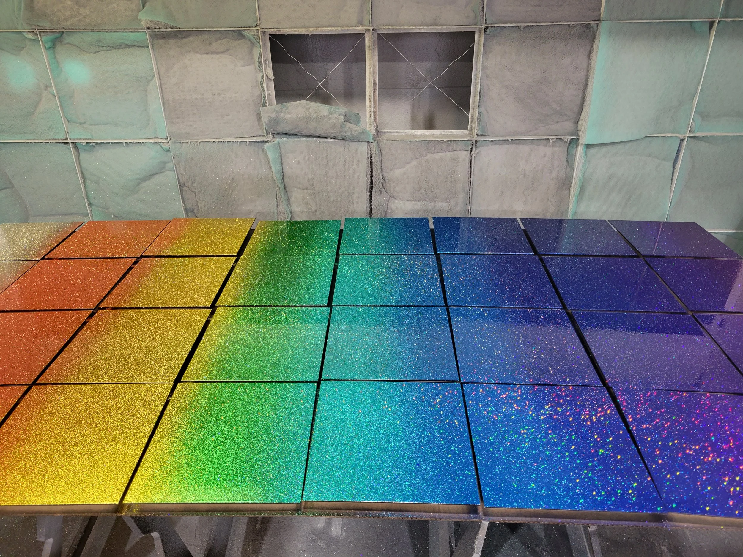 A table with colored glittery tiles arranged in a rainbow gradient from left to right, with a stone wall in the background.
