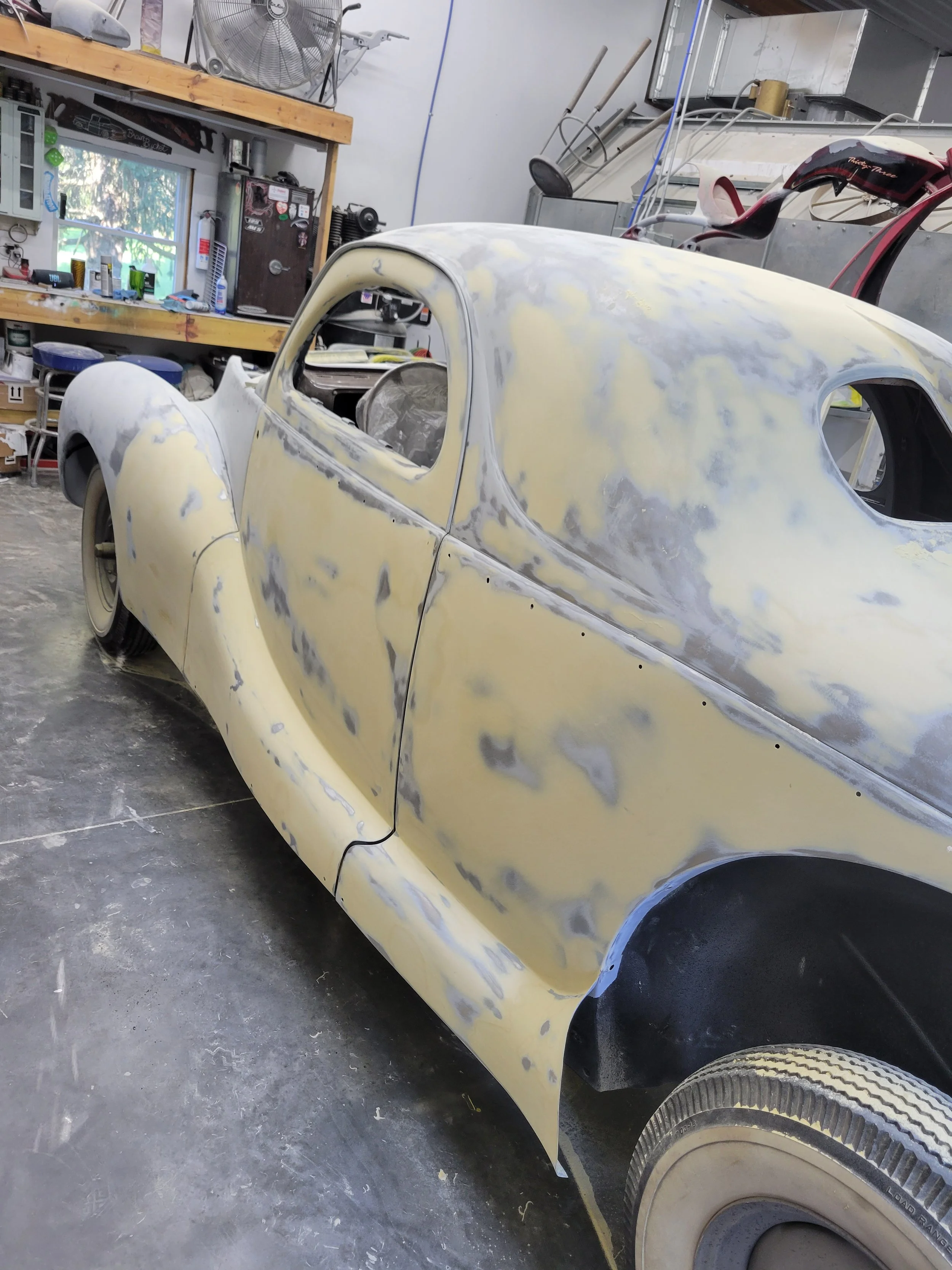 A vintage car undergoing restoration in a workshop, with the body partially sanded and in primer.