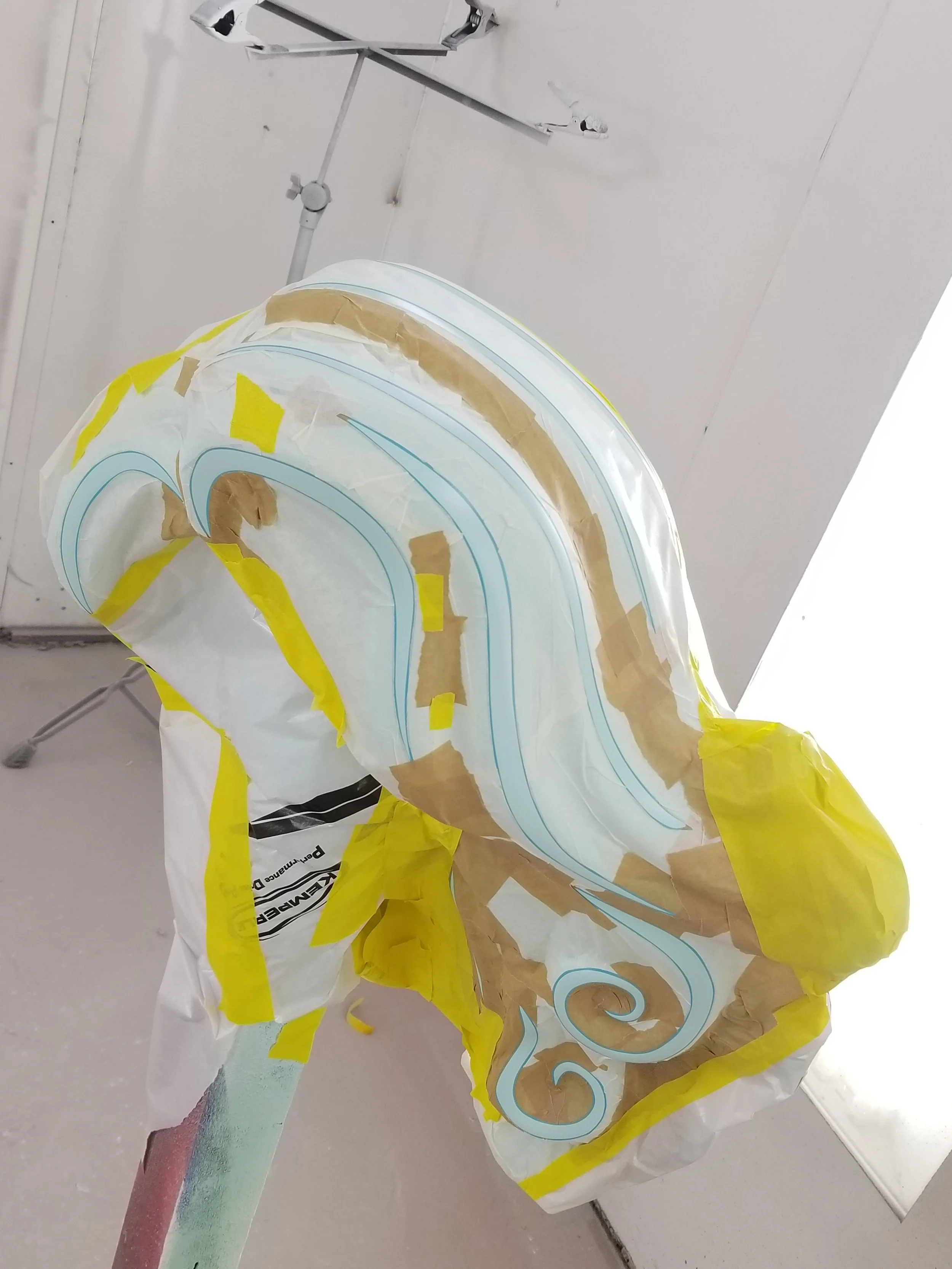 LEGO hair sculpture in a painting booth with masking tape and paper for a paint job, placed on a stand, with a white ceiling and wall in the background.