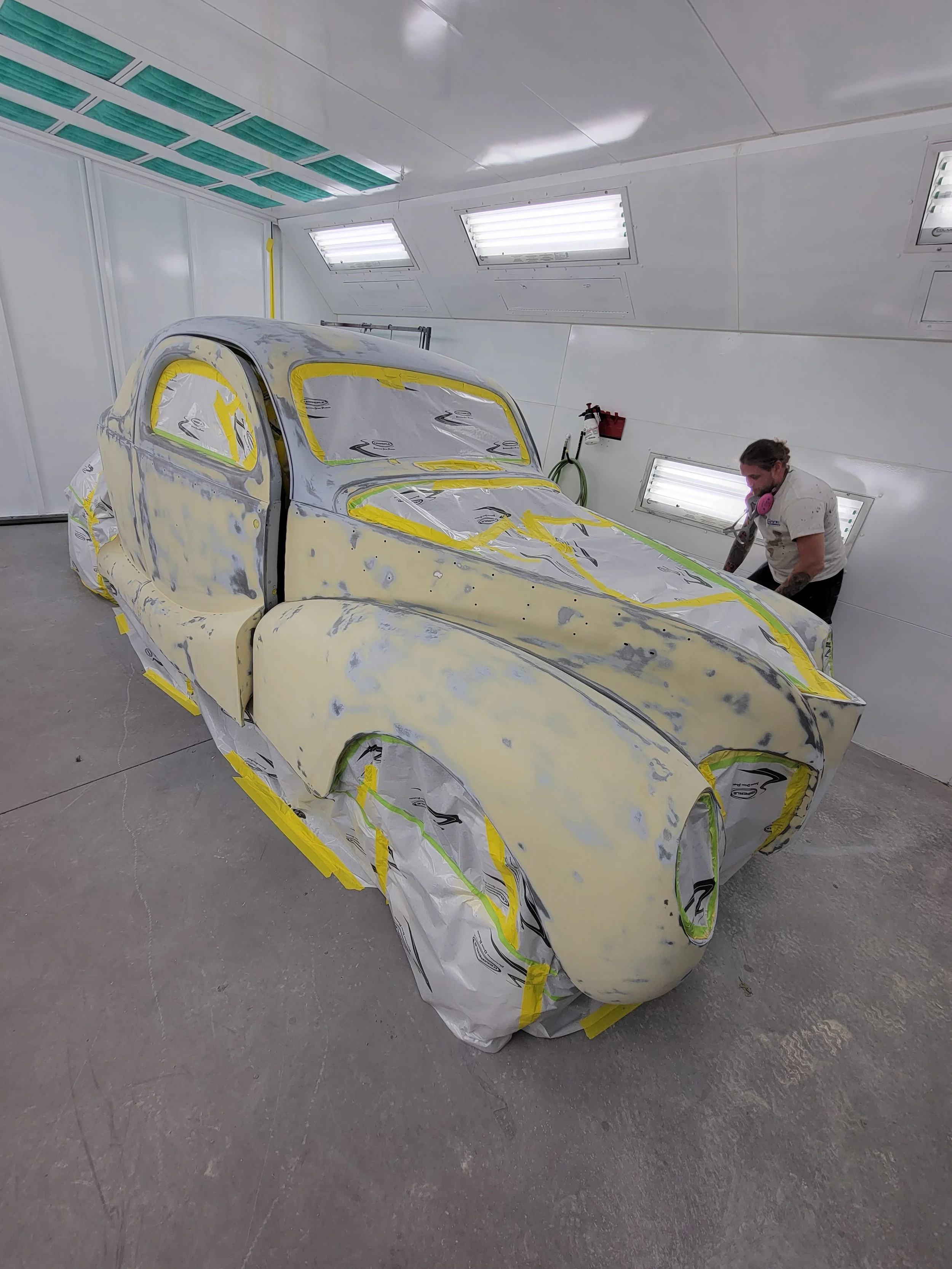 A vintage car is being prepared for painting in a spray booth, covered with masking paper and tape.