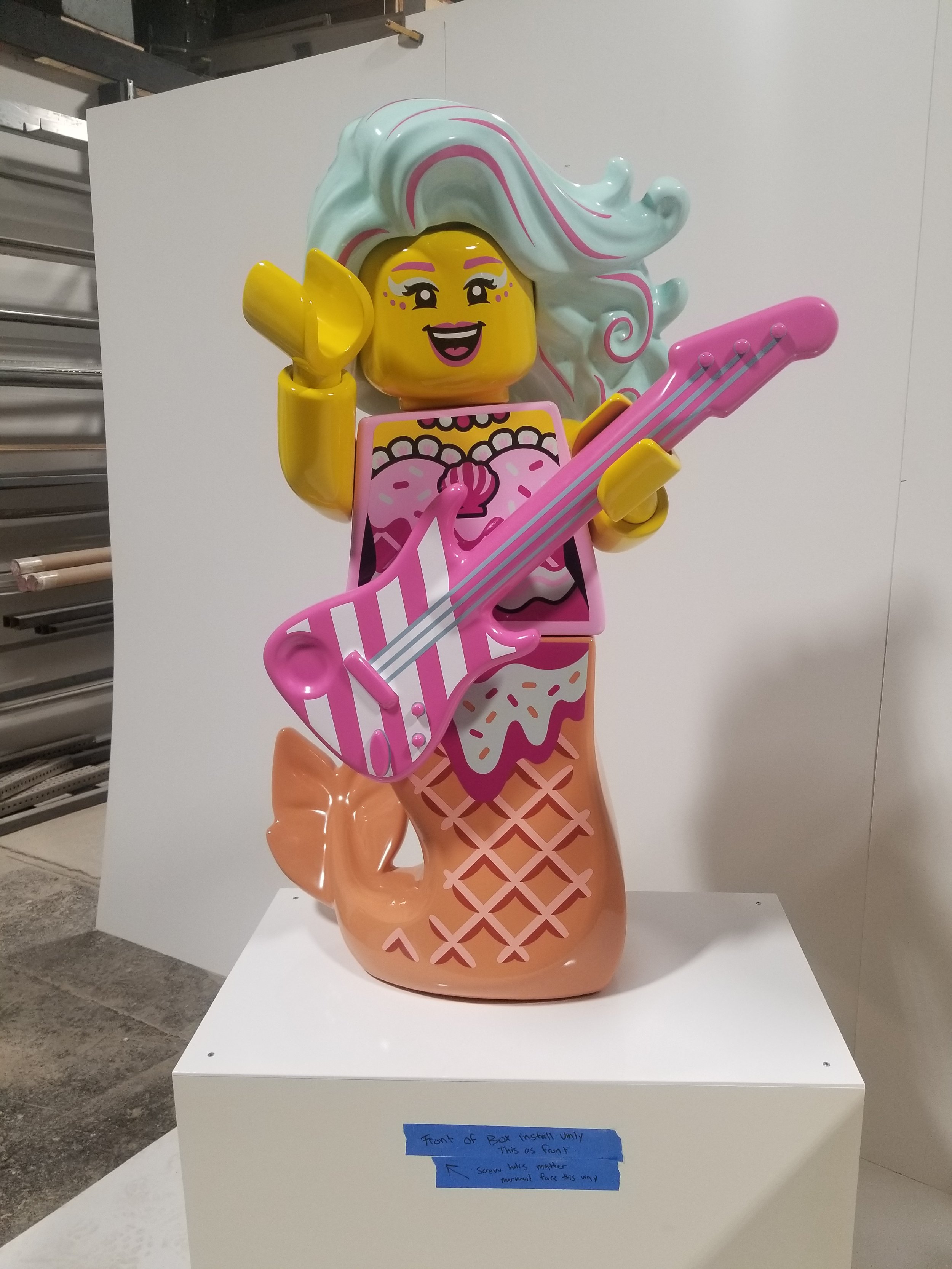 Colorful LEGO sculpture of a mermaid with a long flowing blue and pink hair, holding a pink and white striped electric guitar, and sitting on a large ice cream cone base.