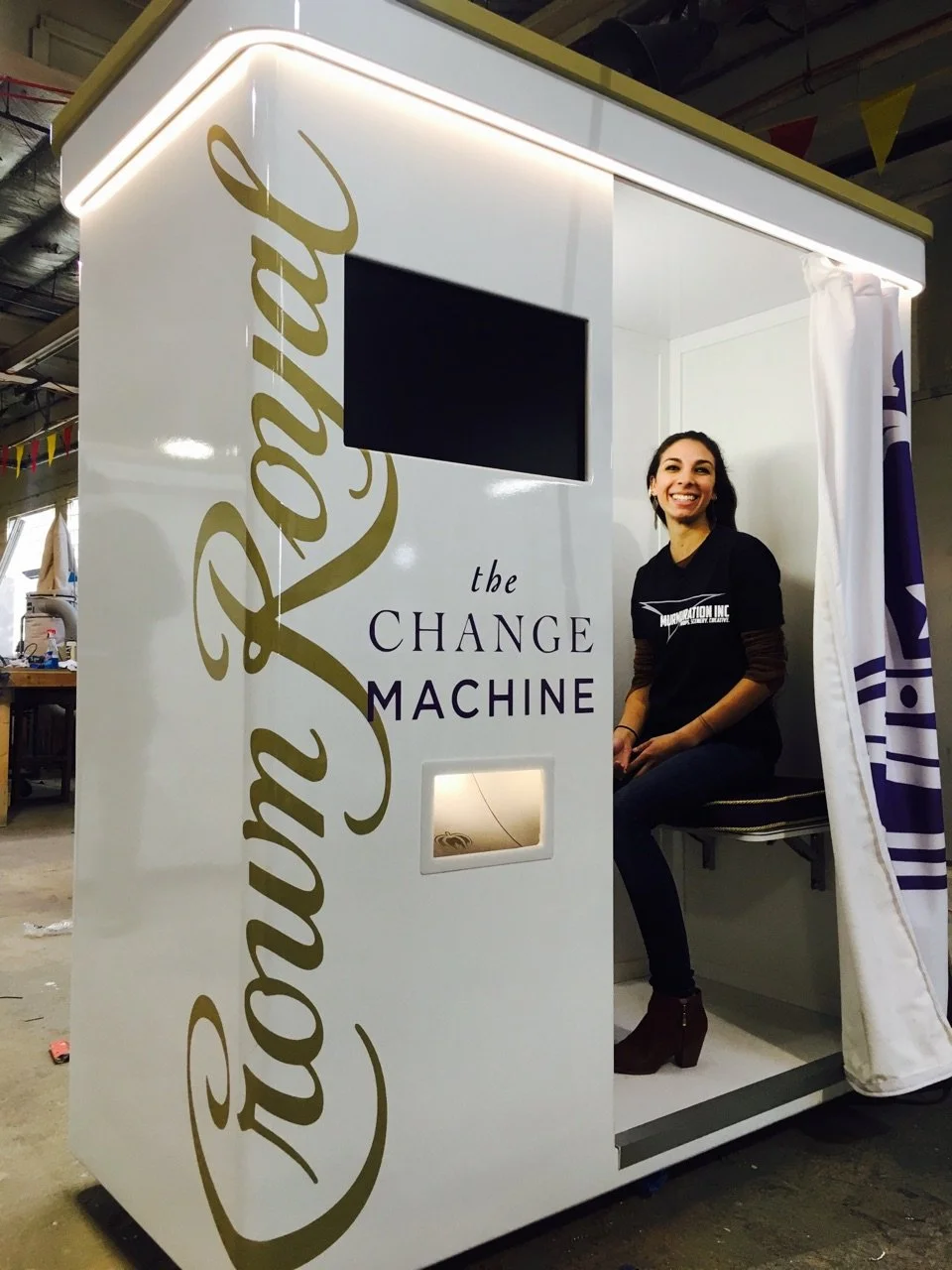 A woman sitting inside a white booth with the text "the Change Machine" and "Your Transformation Inc." on it, with a man smiling.
