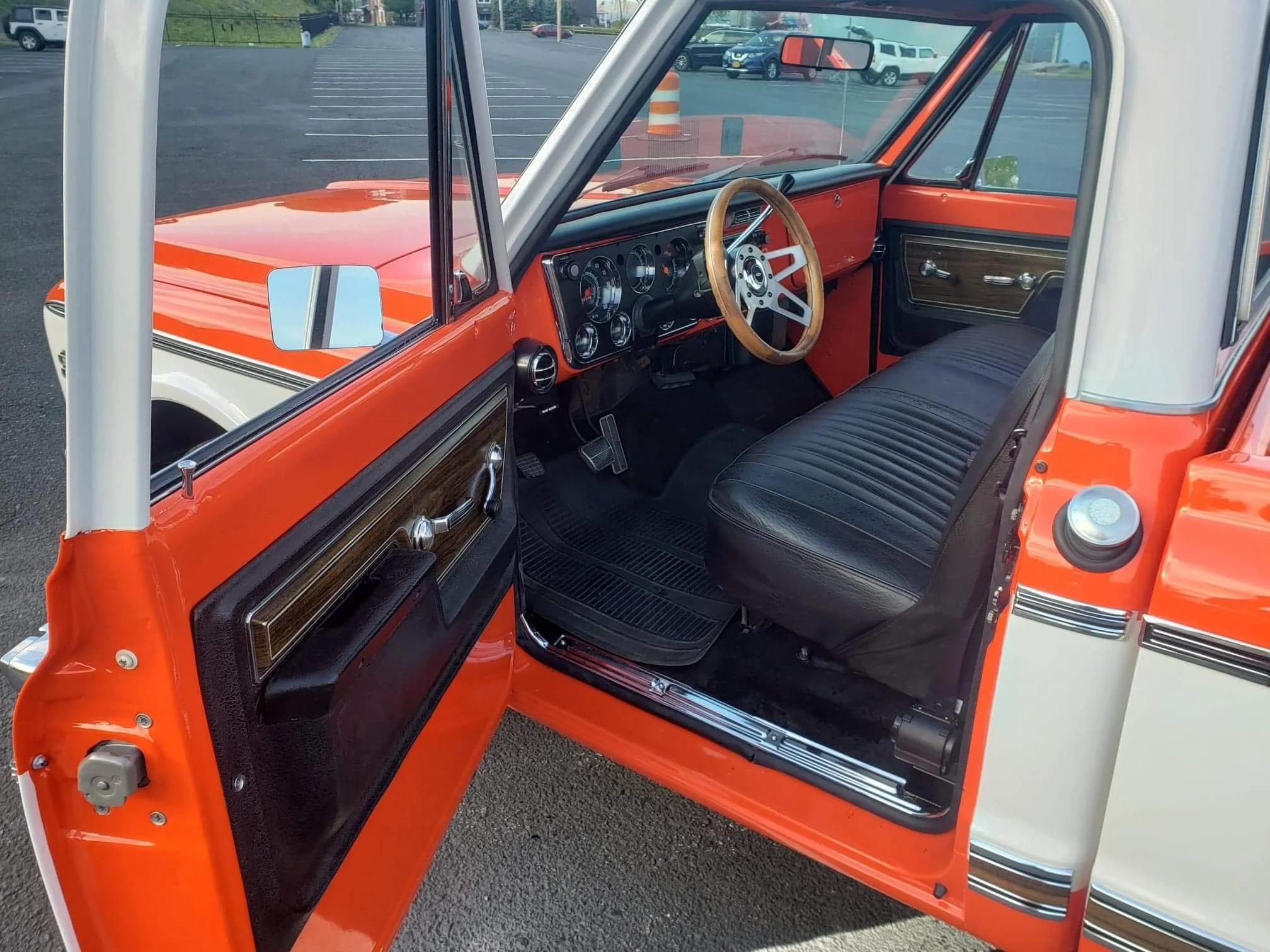 Interior of a vintage orange and white Jeep Wagoneer with a black bench seat, wood panel dashboard, gray steering wheel, and manual window controls, parked in a lot.