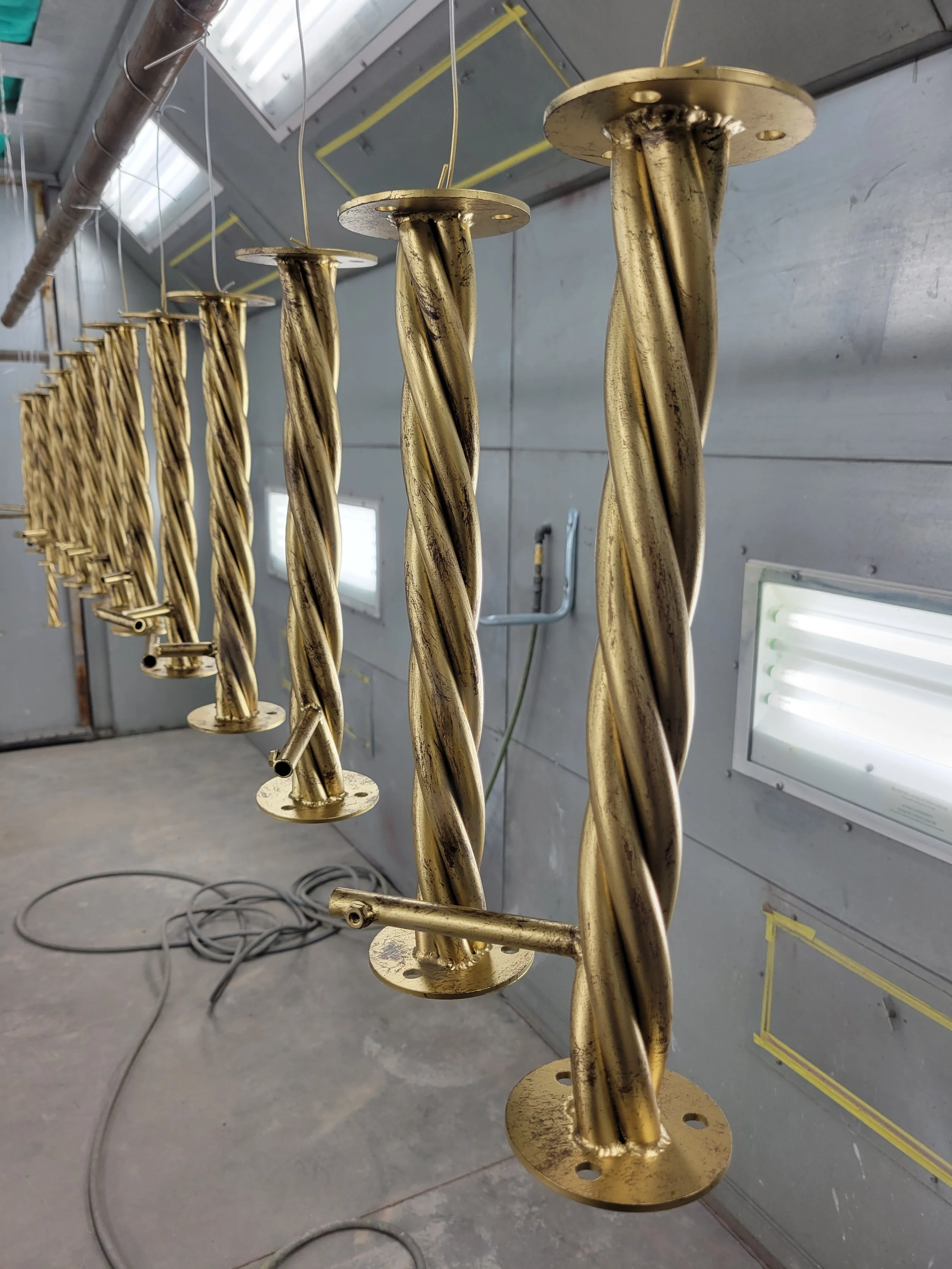 Multiple large, metallic, gold-colored twisted metal rods hanging in a line inside a workshop or industrial space.