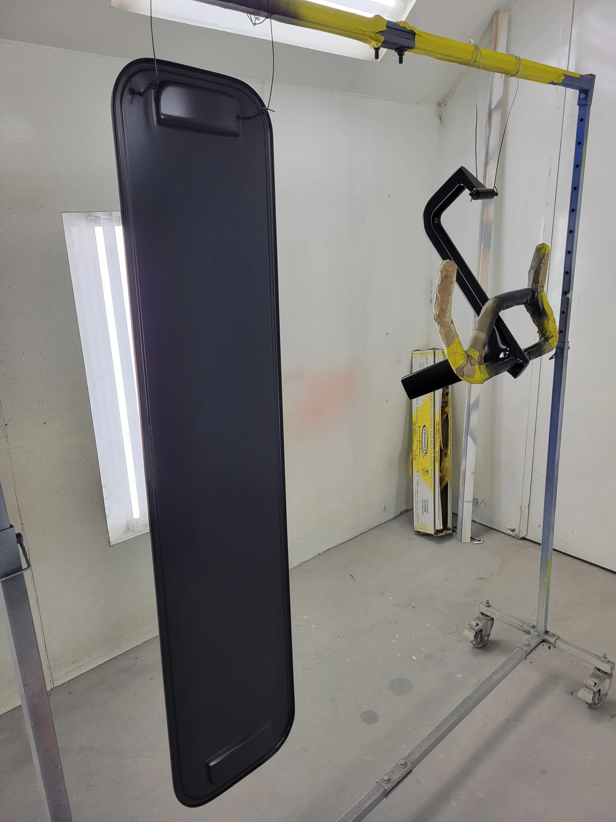 A black metal car side mirror is hanging from a yellow support beam inside a workshop or garage. The mirror is attached to a metal frame with caster wheels, and there are some tools and yellow packaging materials in the background.