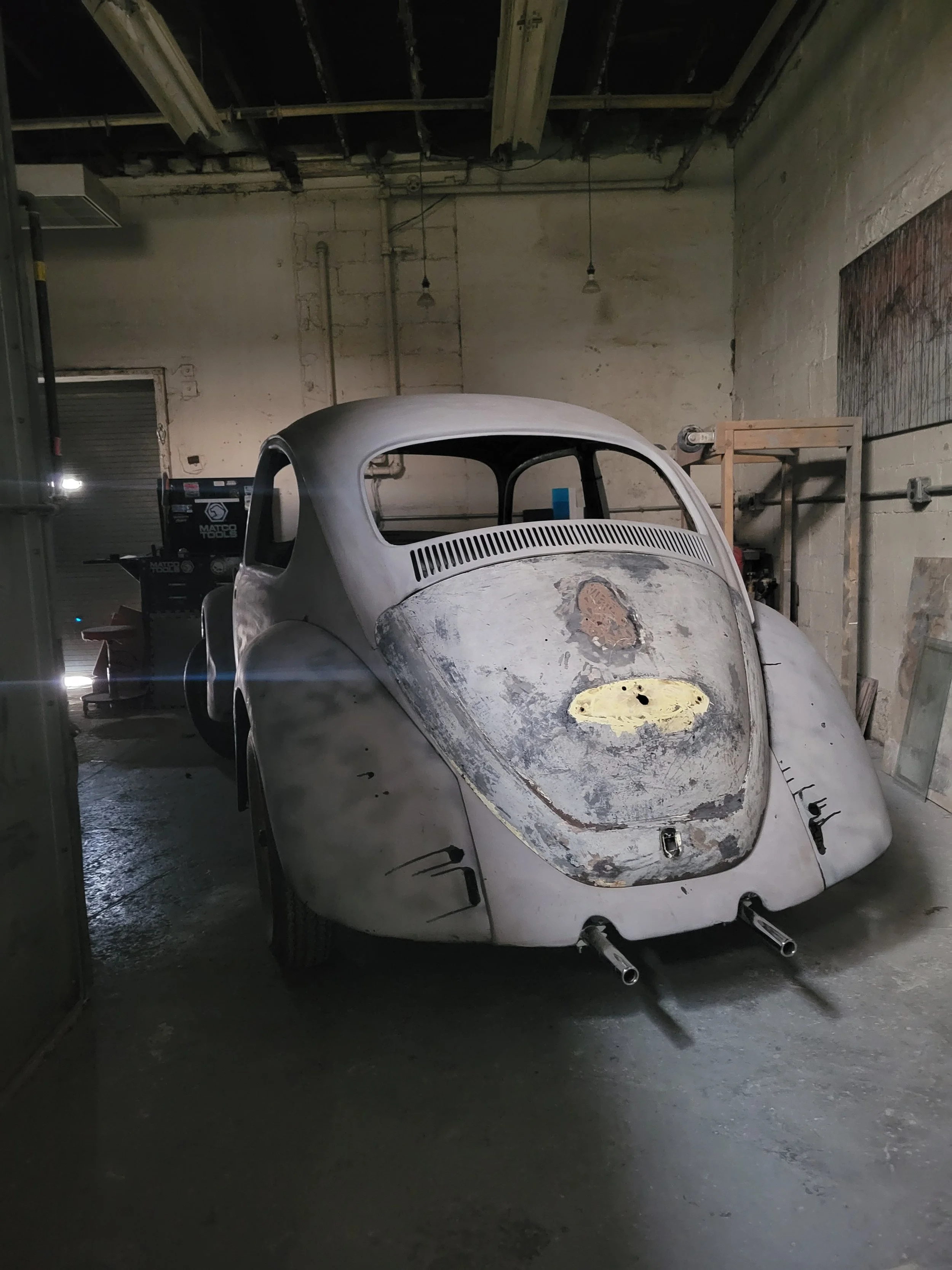An unfinished vintage car with a rounded body in a garage or workshop. The body shows rust and corrosion, and the paint is worn. Exhaust pipes protrude from the rear, and the car lacks windows and a complete exterior.