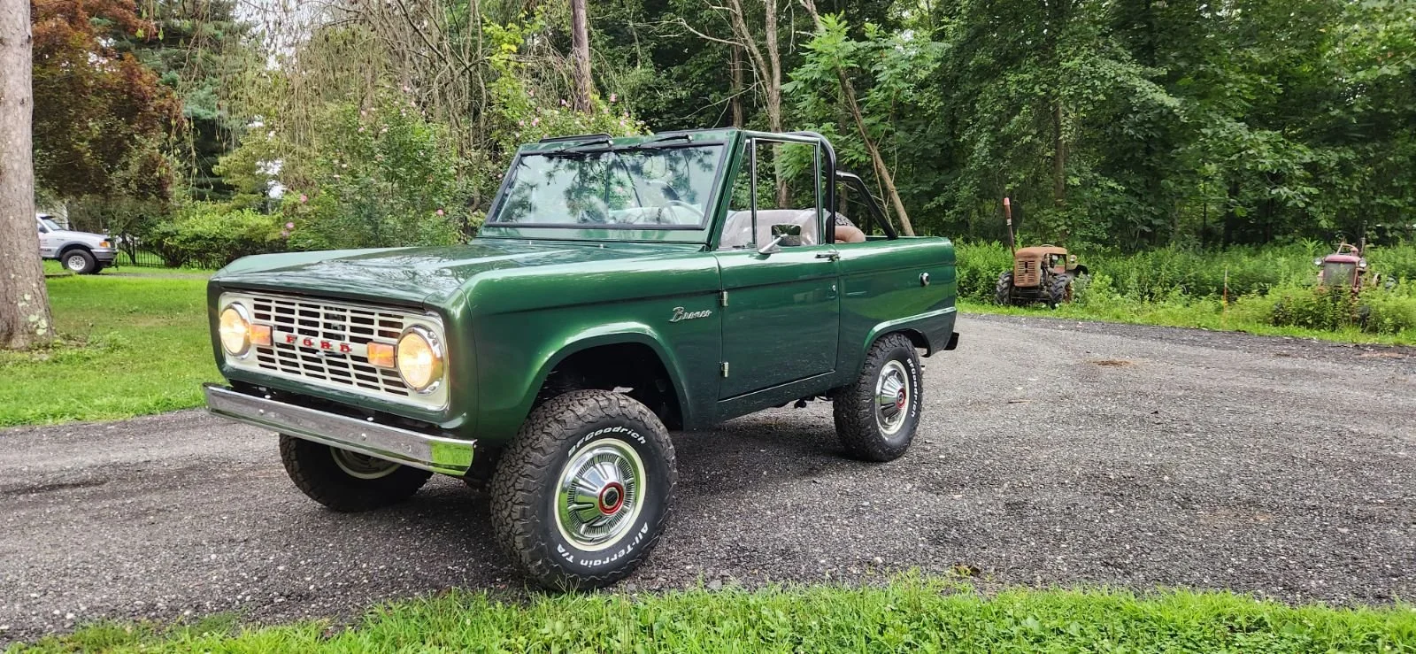 A vintage green convertible car parked on gravel, surrounded by grass and trees, with an old tractor in the background.