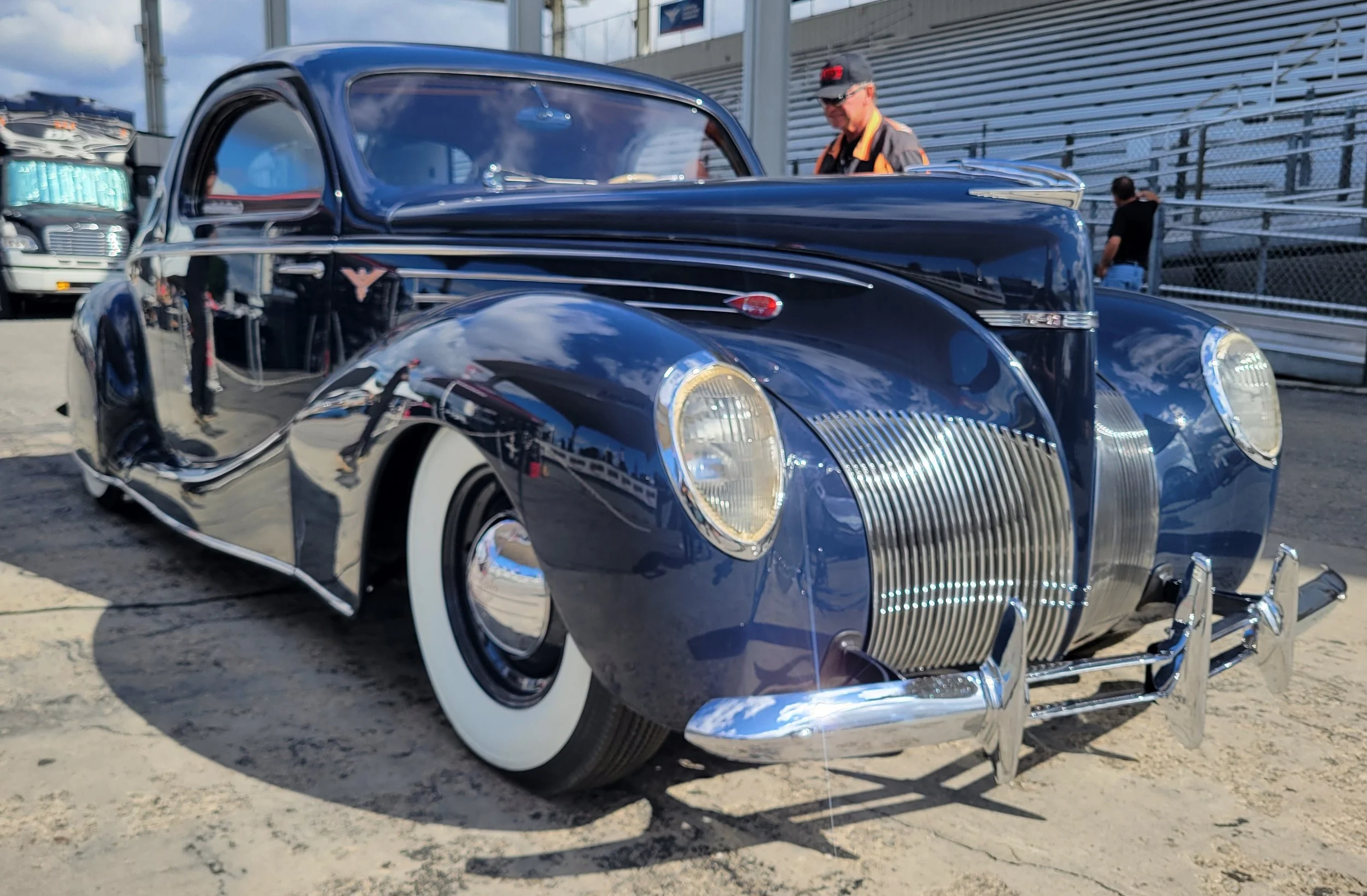 A vintage blue car with chrome accents and white-wall tires parked at an outdoor car show, with two people in the background examining the vehicle.