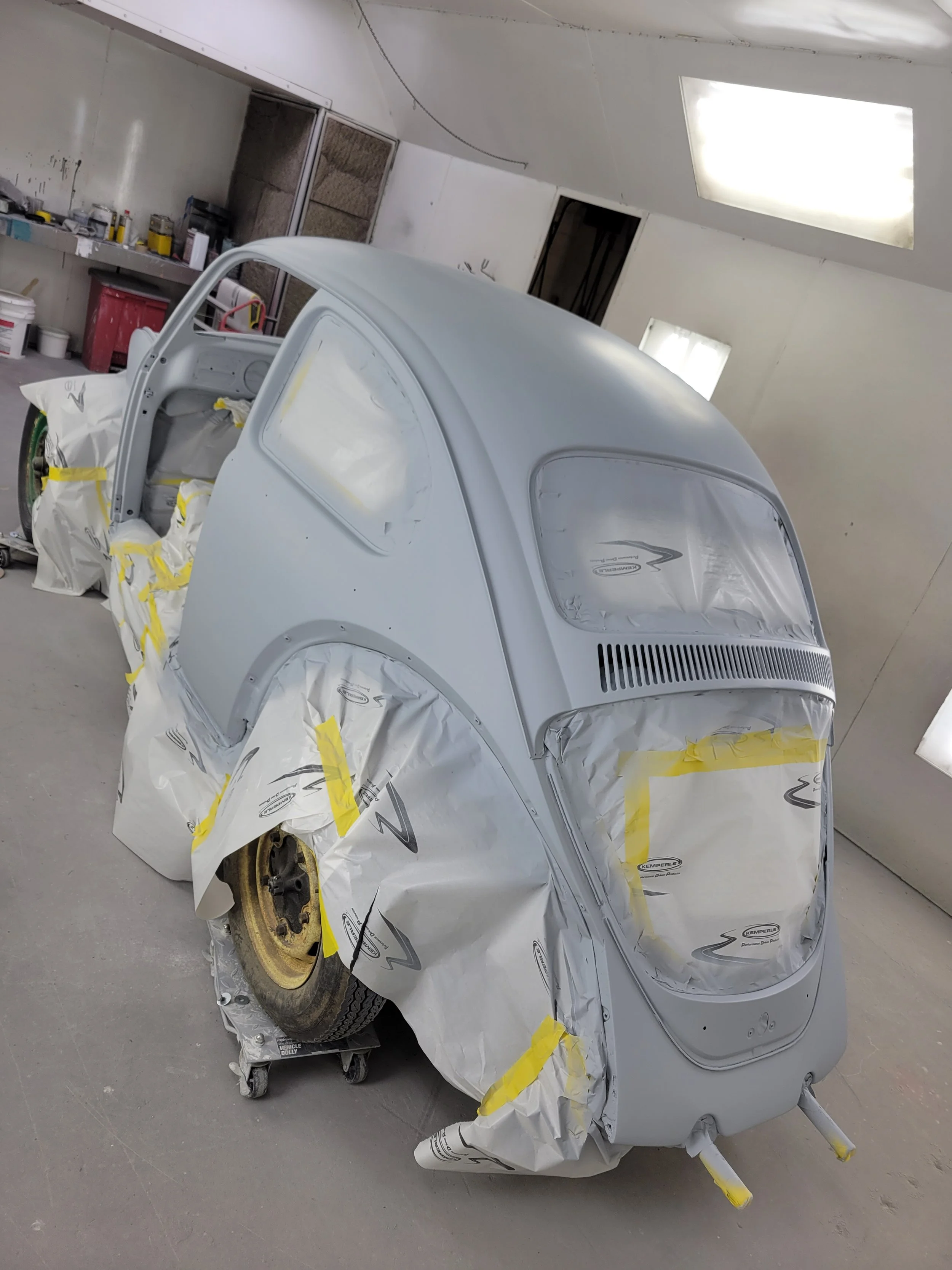 Small vintage vehicle in a workshop being prepared for painting, covered with masking paper and tape, with a spare tire mounted on the side.