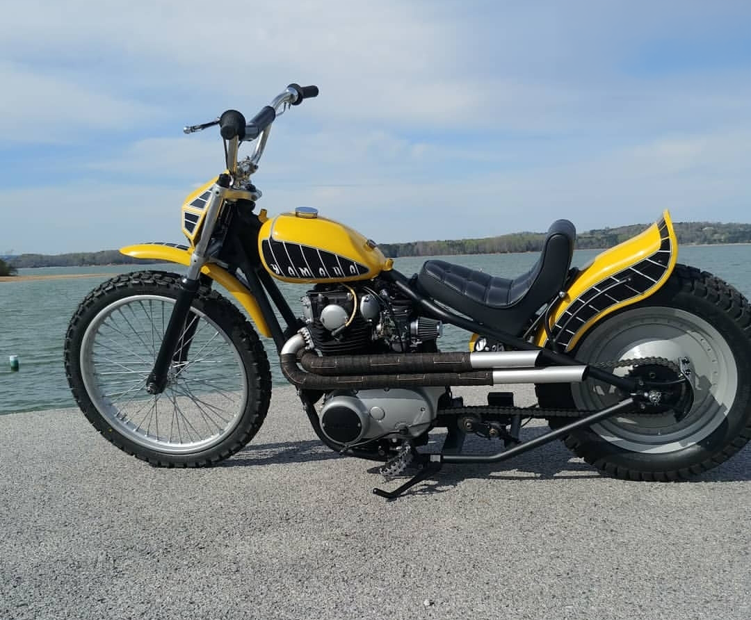 Yellow vintage motorcycle with black accents parked on a concrete surface near a body of water under a partly cloudy sky.