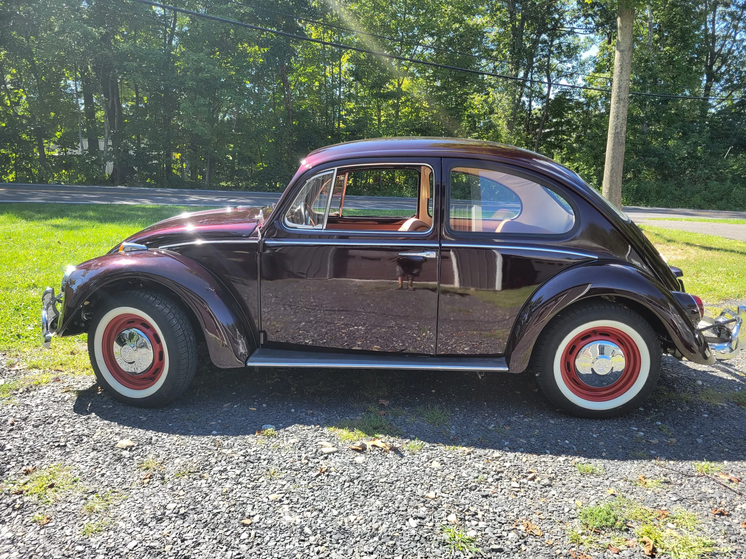 A vintage black Volkswagen Beetle car parked on a gravel surface with green grass and trees in the background.