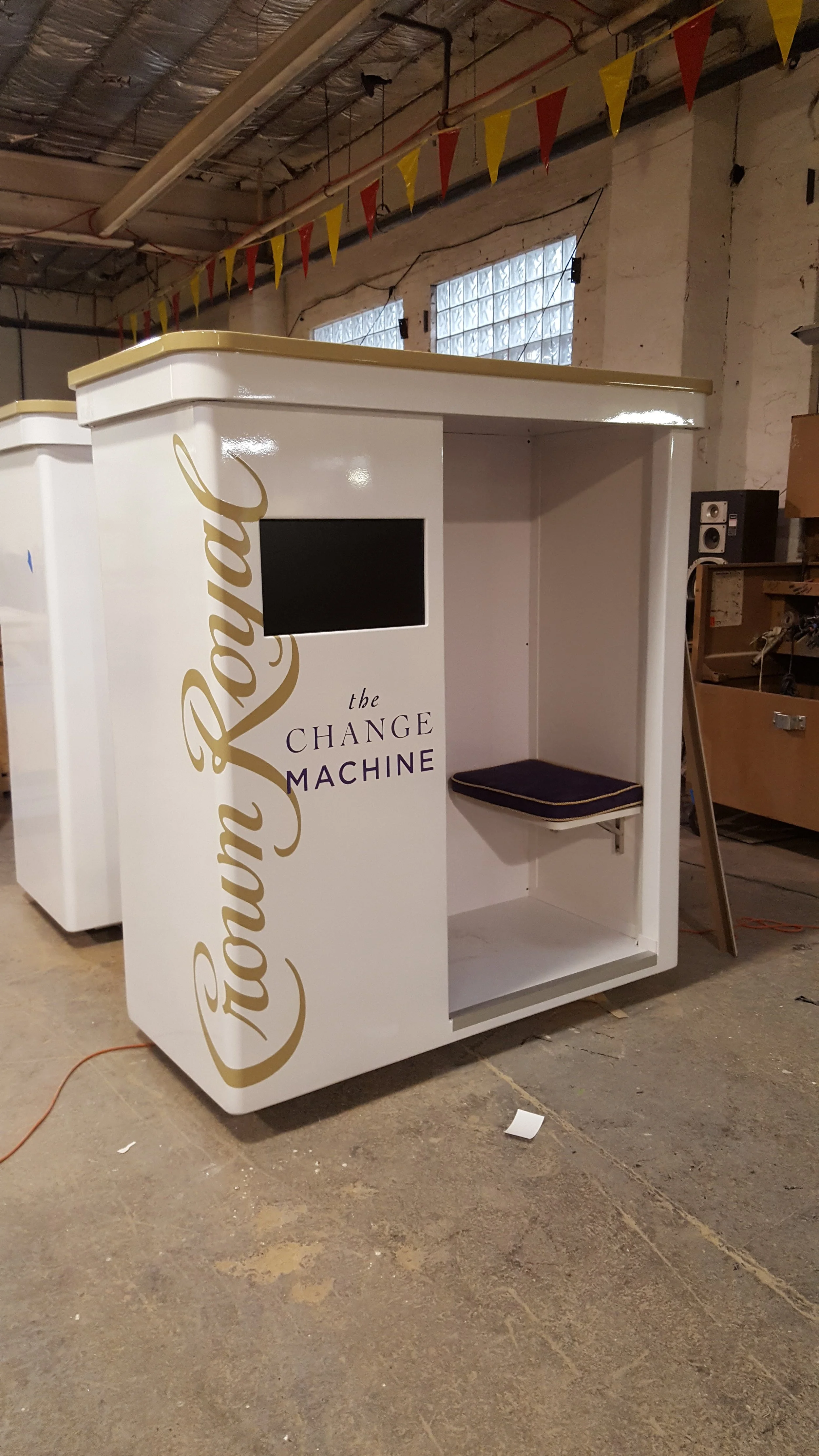 A white photo booth with gold lettering that reads 'the CHANGE MACHINE'. It has a small screen and a cushioned seat inside, with a large open compartment at the bottom. The photo booth is situated in an industrial-style room with exposed brick walls,