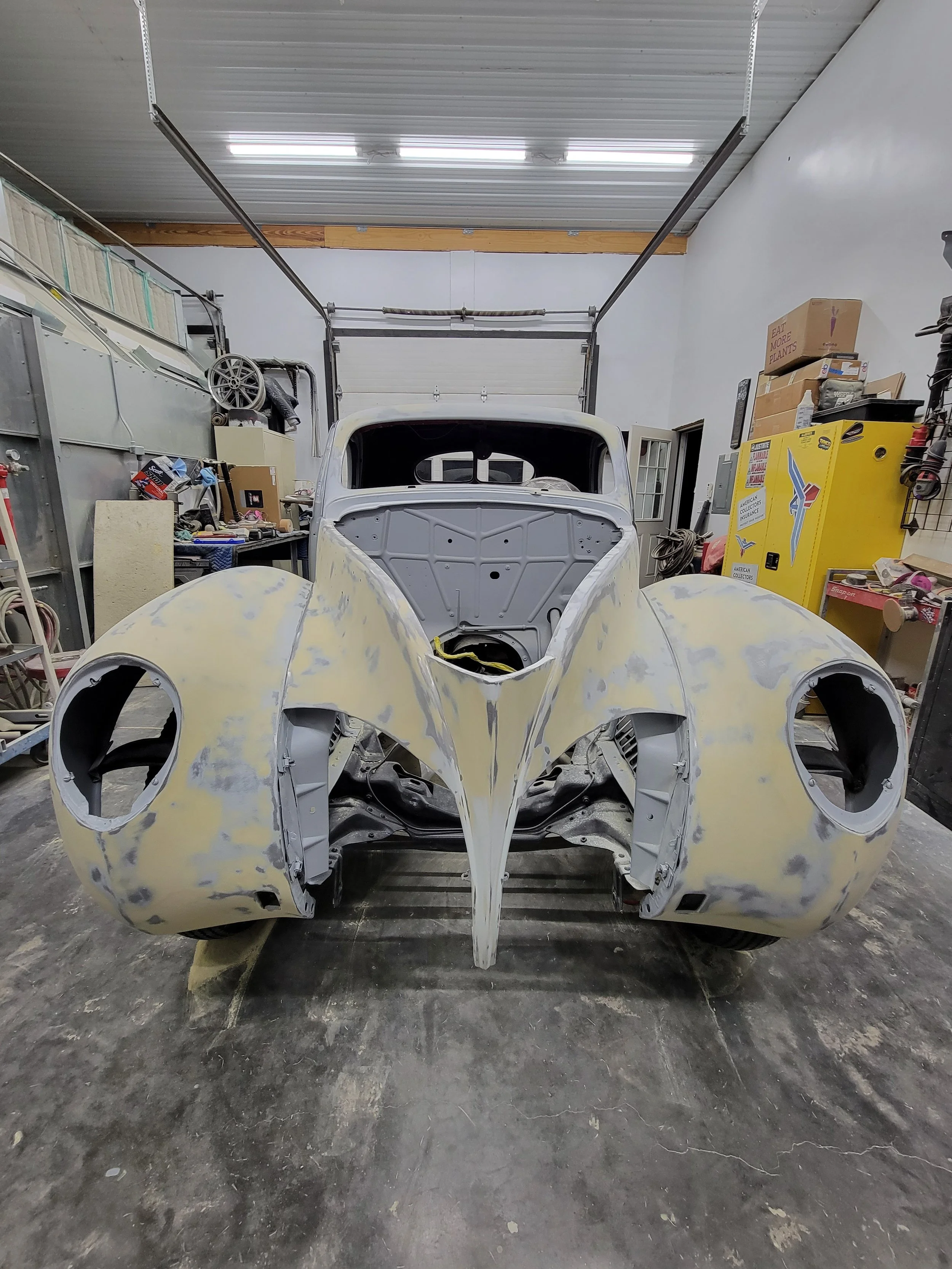 A vintage car body under restoration in a workshop, with a garage door and tools in the background.