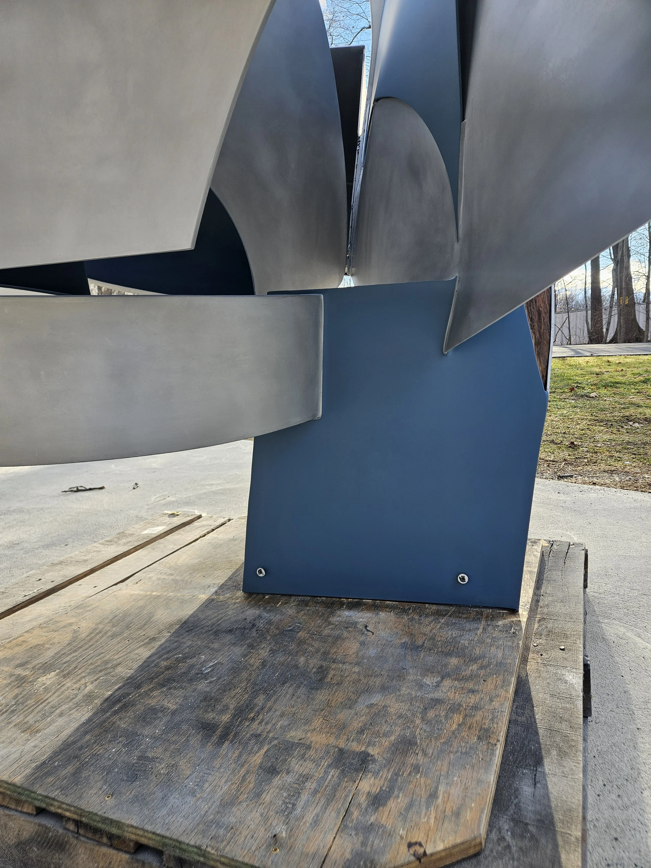 Close-up of a modern abstract metal sculpture with blue and silver pieces, mounted on a wooden base outdoors with trees and sky in the background.