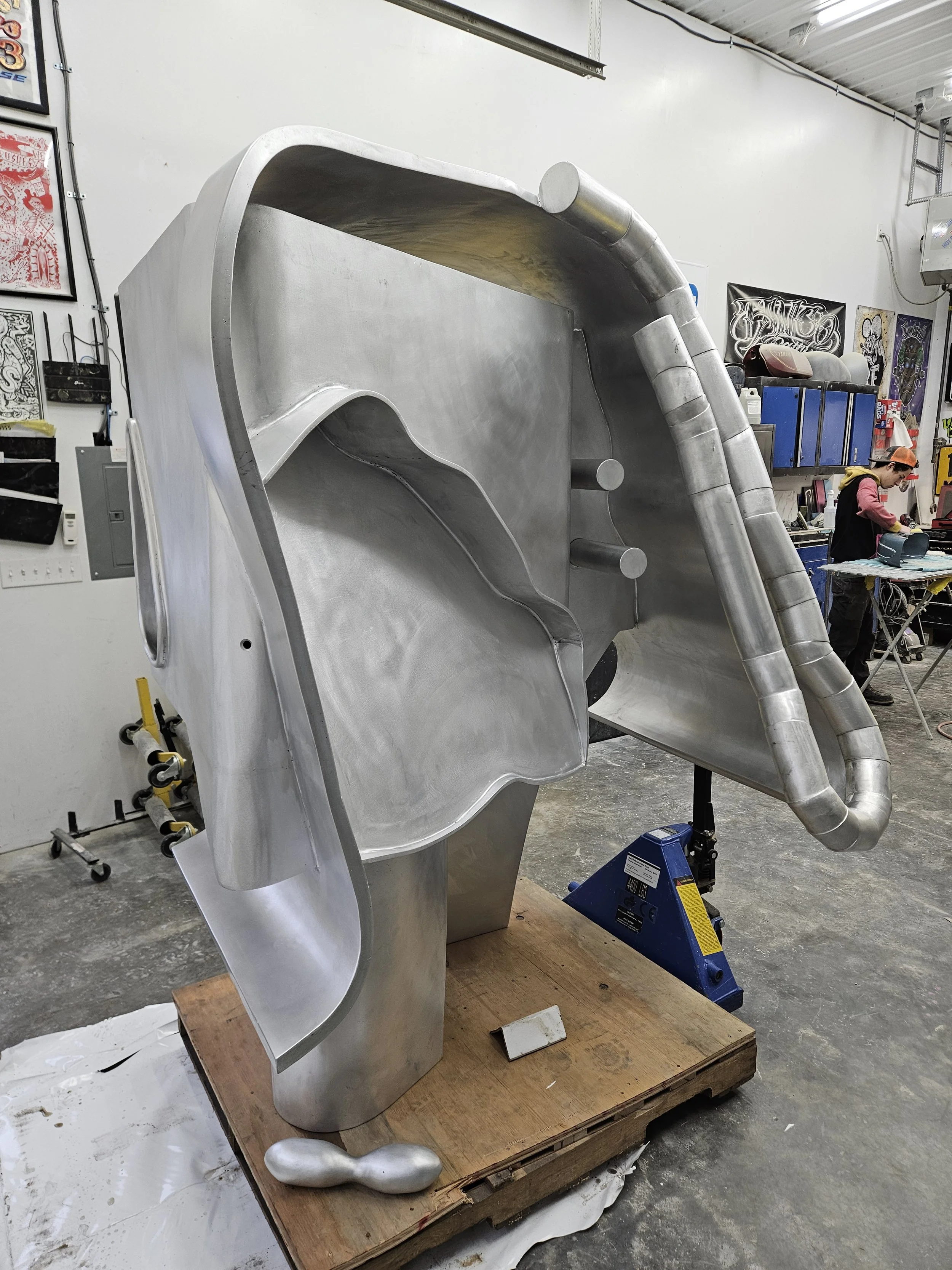 Large metallic sculpture or structure with abstract, flowing shapes, positioned in an industrial workshop with tools, storage cabinets, and a person working in the background.