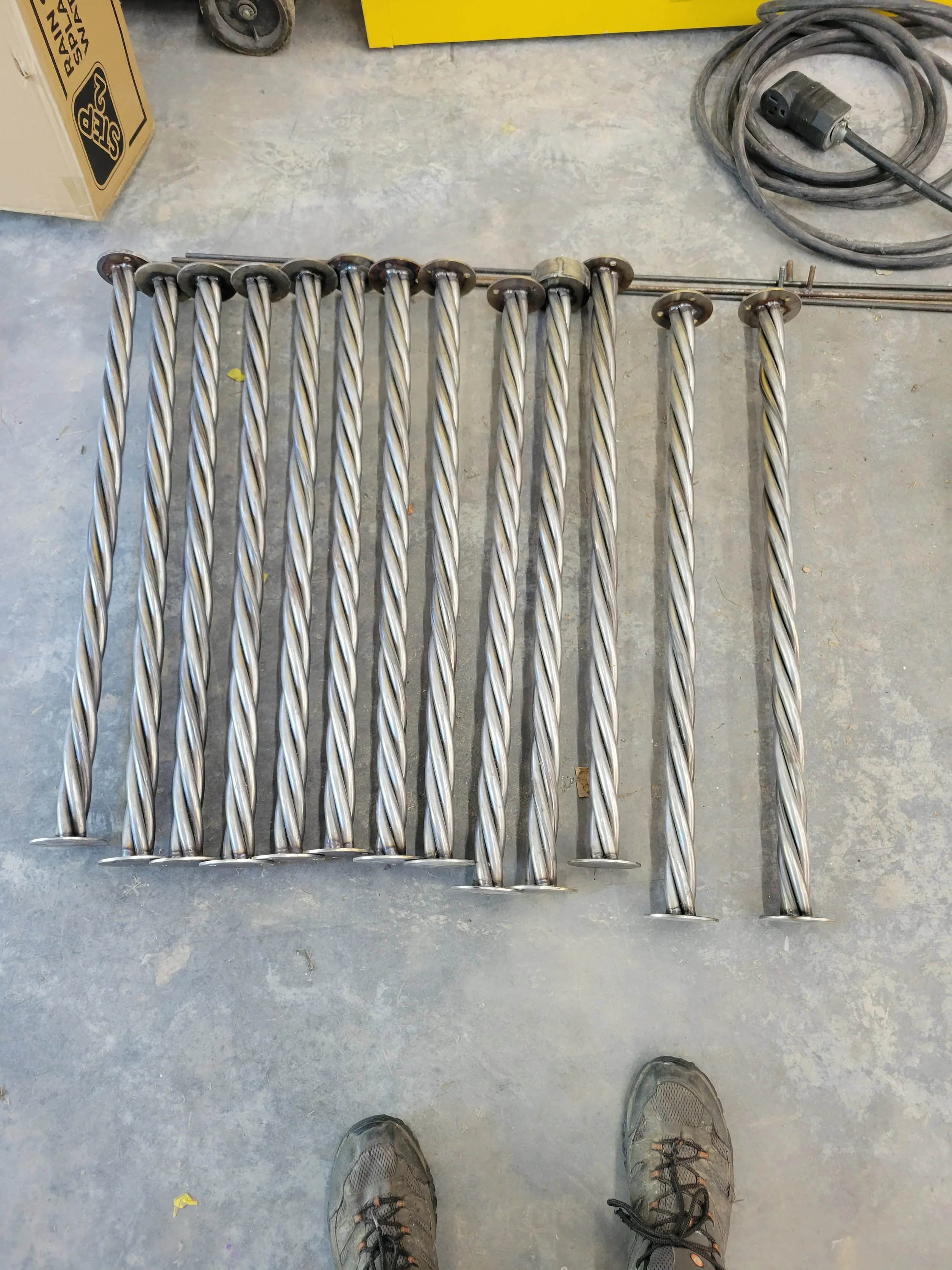 A set of metal twisted rod bars laid out on a concrete floor in a workshop or garage. A person wearing gray shoes is taking the photo.