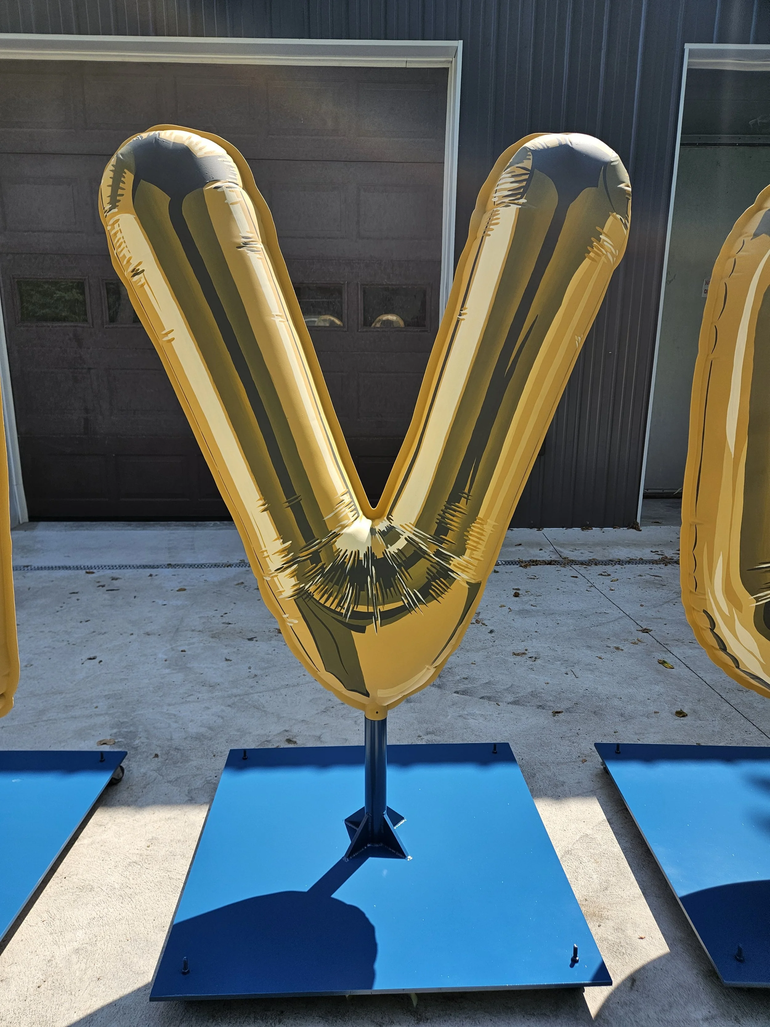 Large yellow inflatable balloon shaped like the letter V, mounted on a blue stand outdoors in front of a dark garage door.
